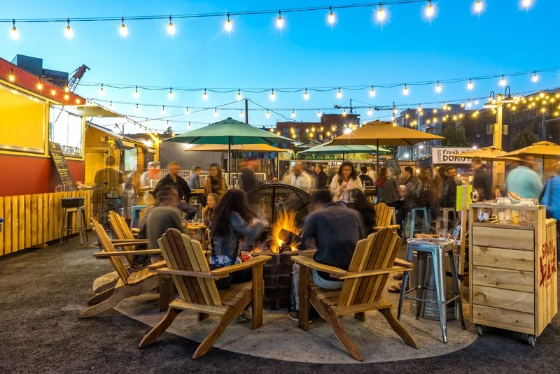 People gather to eat around another fire pit in custom cedar lawn chairs. In the background, the food truck park is bustling.