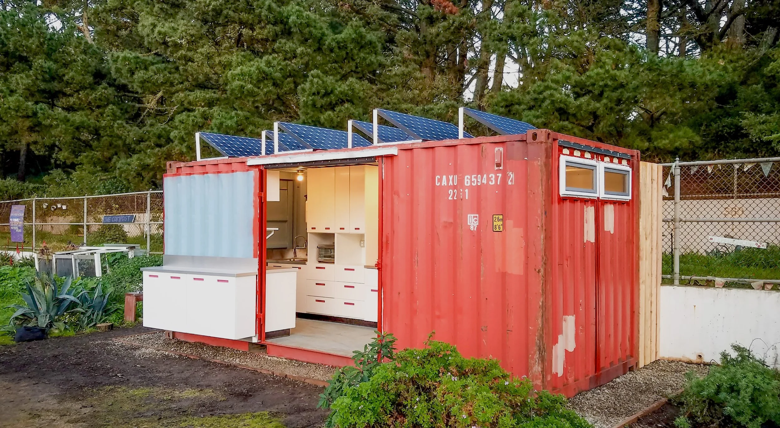 A shipping container converted into an outdoor kitchen, situated in a garden. Solar panels are mounted on top, and the door opens to reveal custom cabinetry and countertops.
