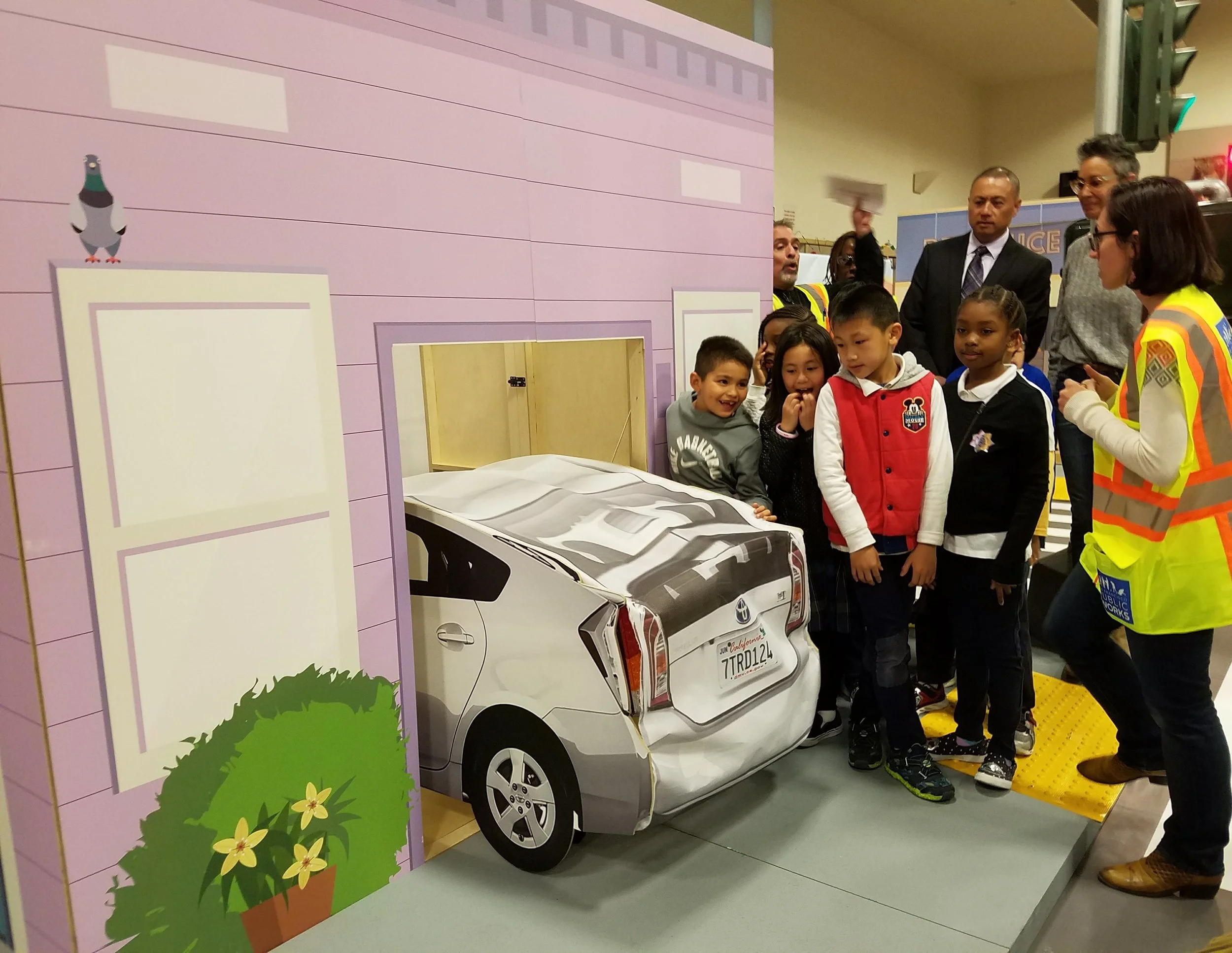 A group of children and adults gather around a model car backing out, teaching them to be on-alert, even when walking on the sidewalk.