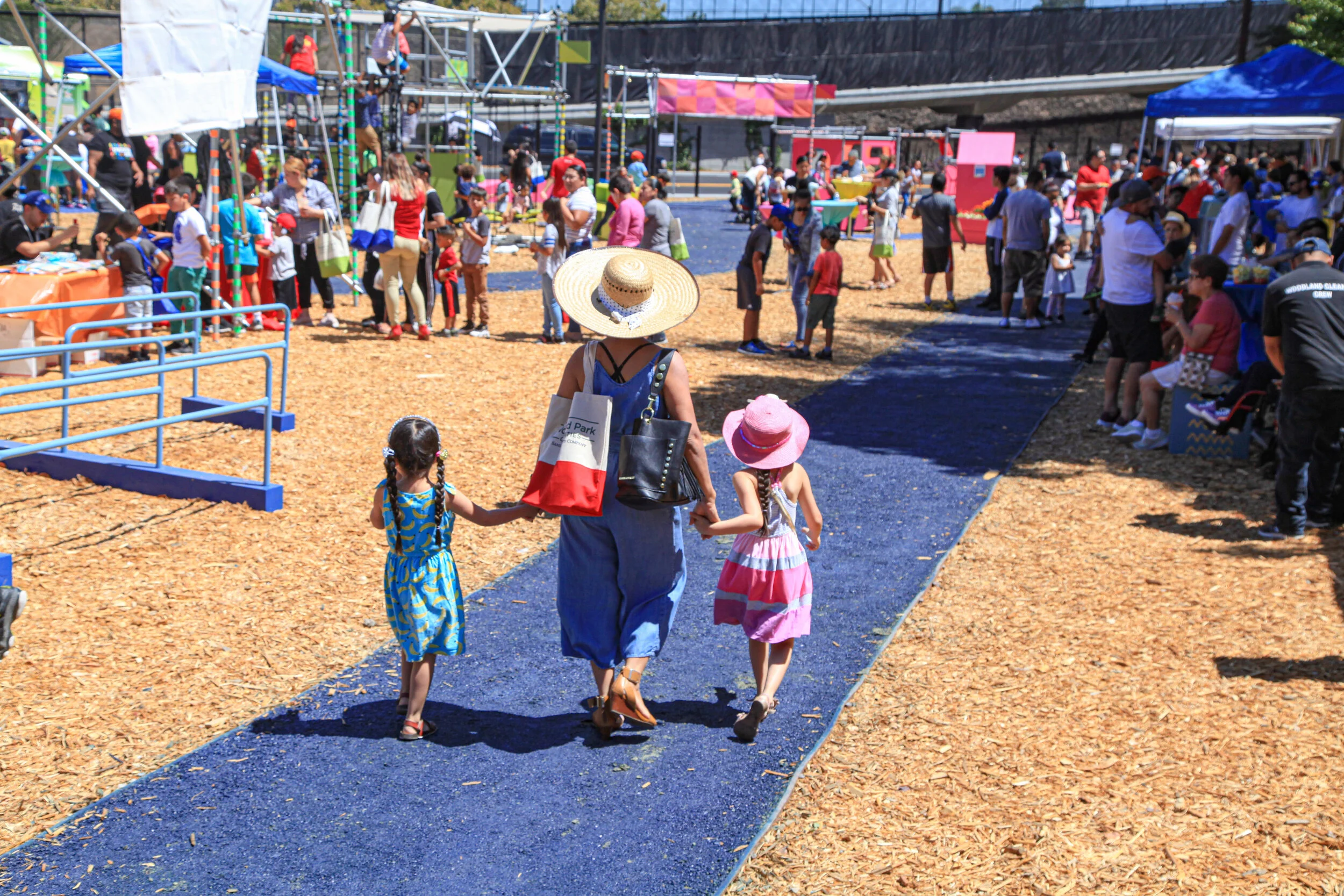 Two girls hold their mothers' hand as they walk along the path towards a series of bright playstructures. Children and adults are all having a blast!