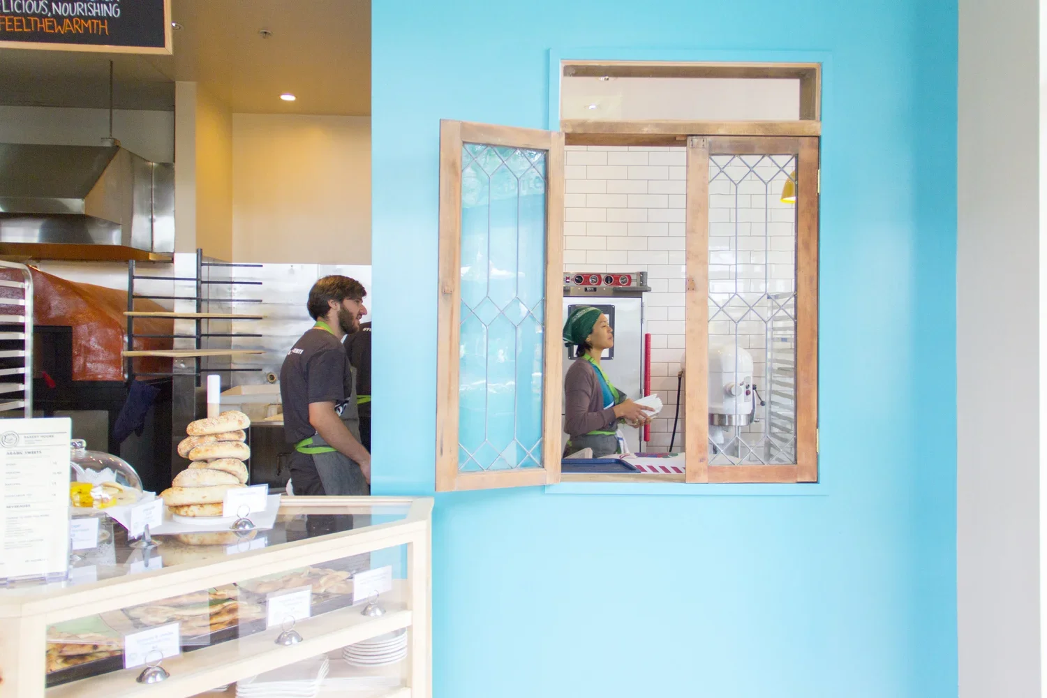 A display case of baked goods is in the foreground, with an ornate rustic and partially open window revealing the innerworkings of the bakery behind it.