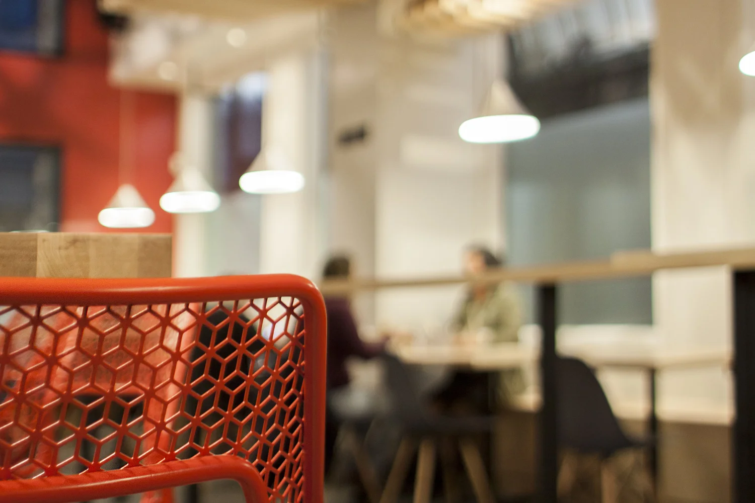 Close-up of a red metal chair in a modern cafe with blurred background of people sitting and white hanging lights.