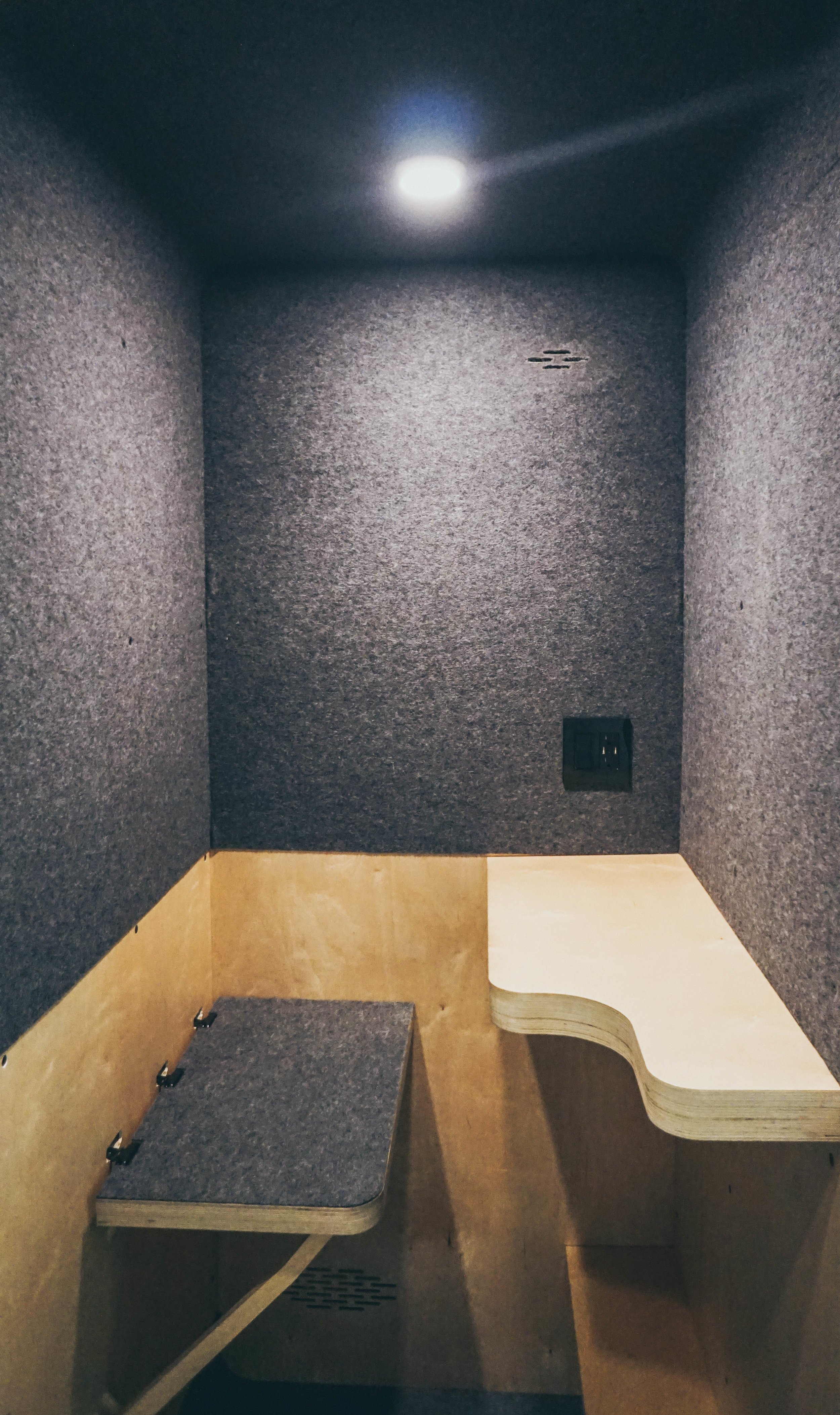 Small enclosed space with gray carpeted walls and a wooden seat and backrest, with a power outlet on the wall.
