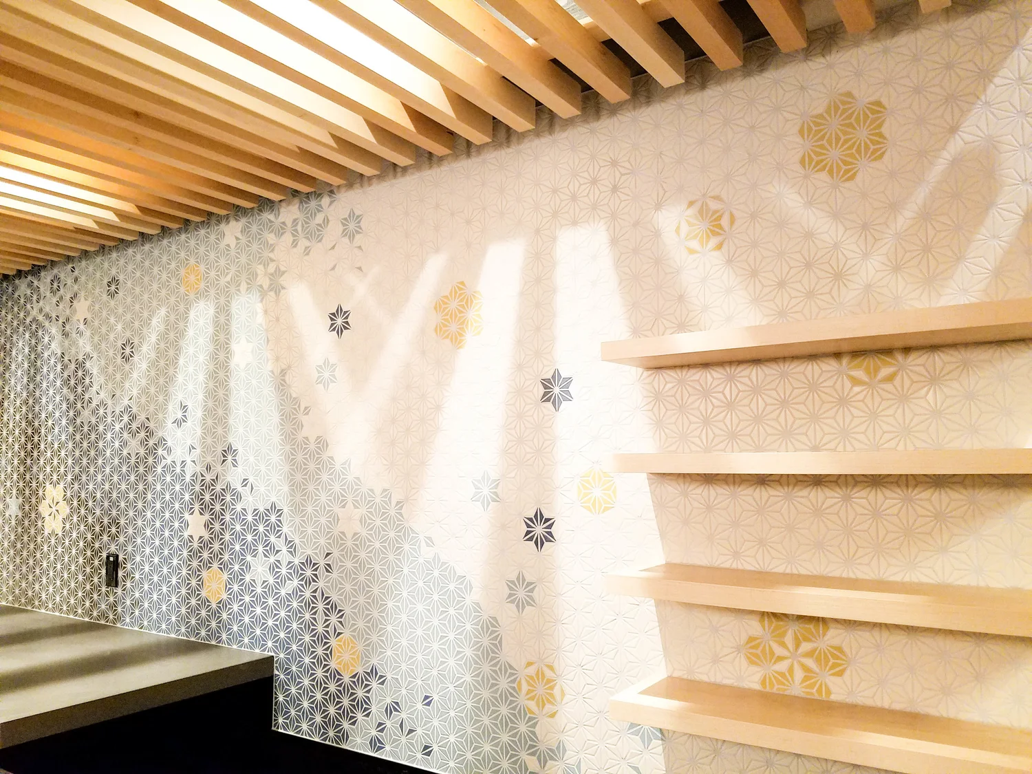 Interior wall with geometric patterned wallpaper and floating wooden shelves, with ceiling slats.