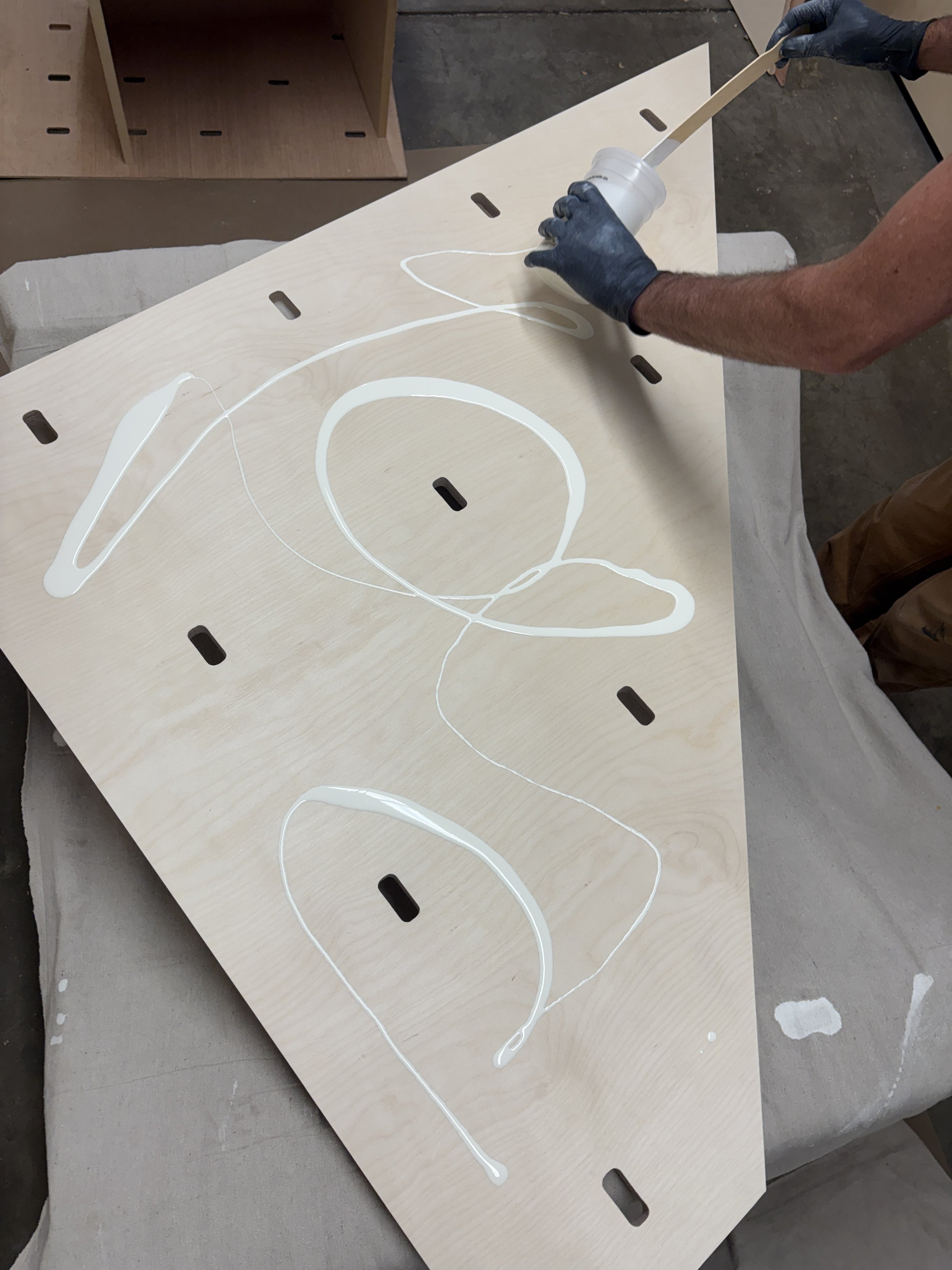 A person wearing black gloves is applying white liquid paint or glue on a wooden surface with a brush or spreading tool. The surface has pre-cut slots and is placed on a padded object for support.