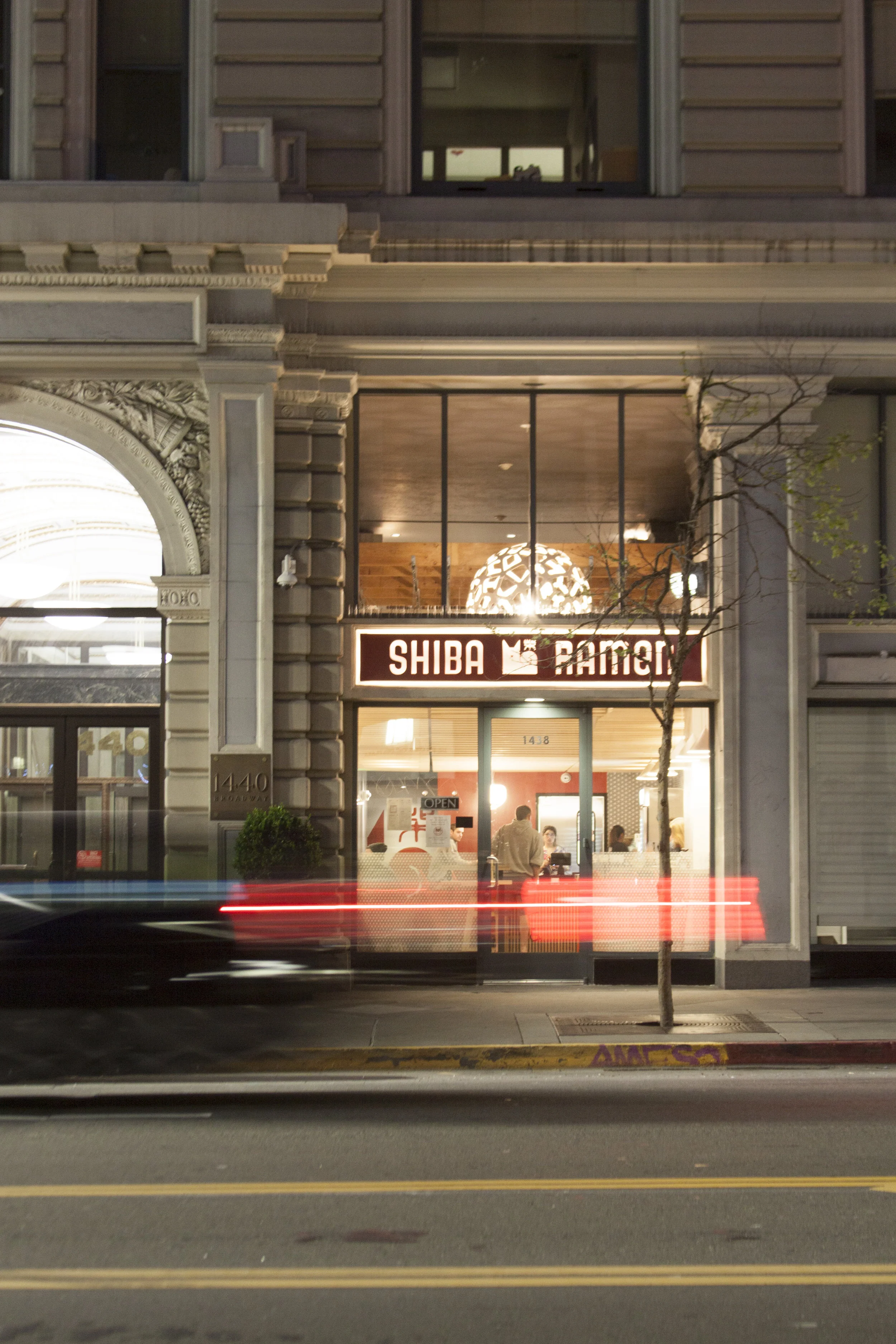 Front entrance, with an entire glass facade and custom back-lit sign, with a car in front captured by motion blur.
