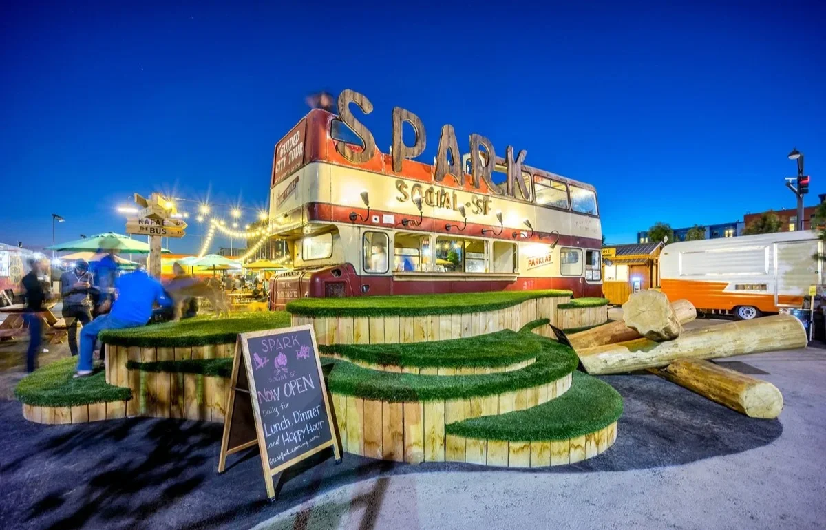 A tiered seating moment and stack of logs invite visistors into Spark Social, offering places for kids and dogs to play. In the background, the food truck park is bustling with patrons under string lights and an evening sky.