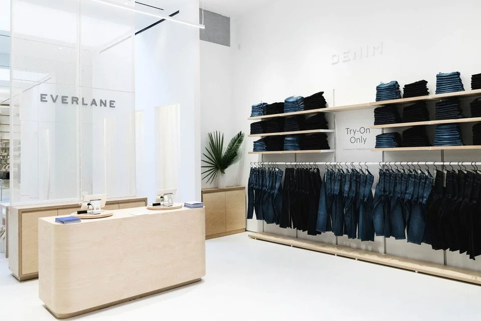 Clothing store interior with shelves of folded jeans and hanging pants, a white counter with point of sale terminals, and a decorative palm plant.