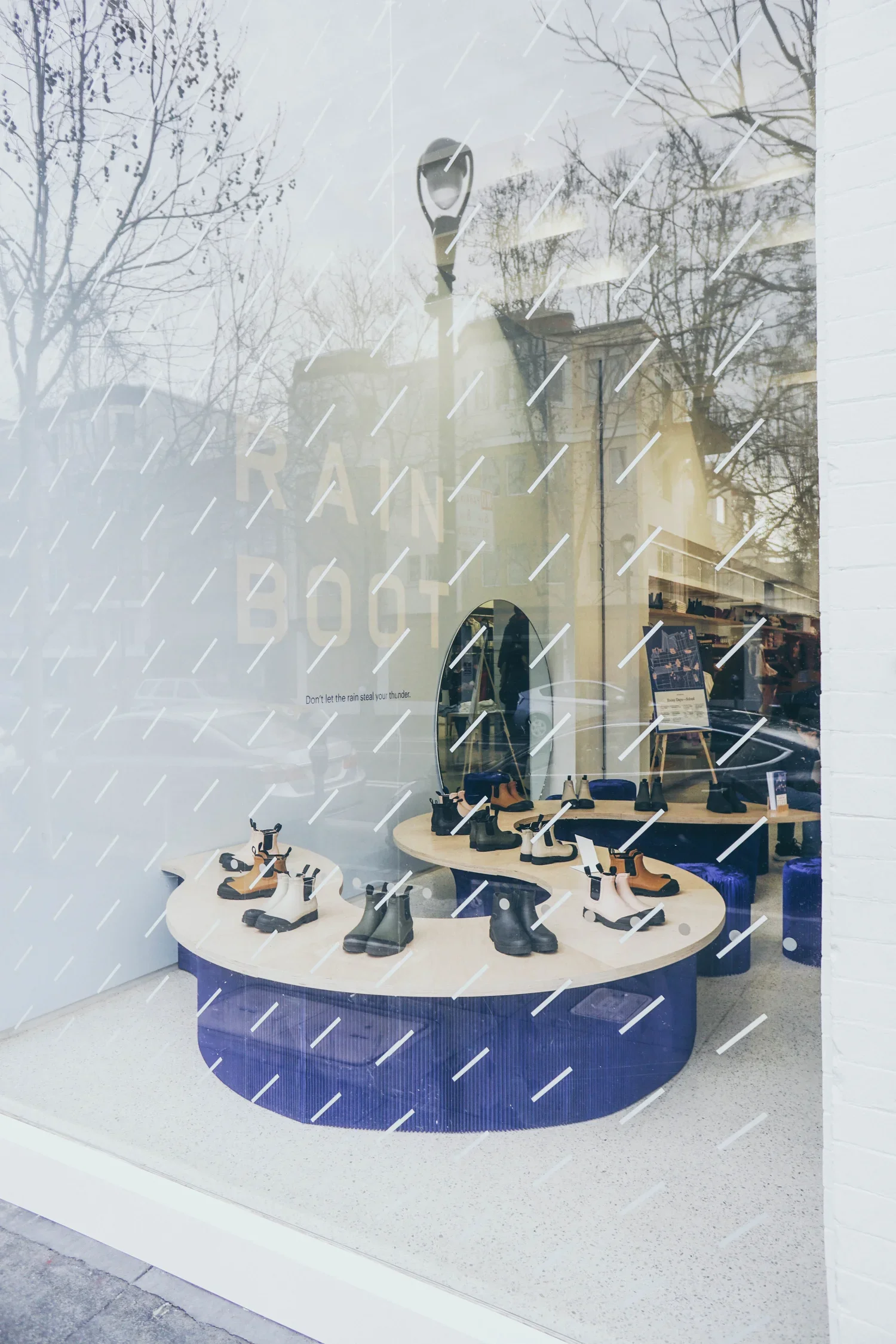Store window display featuring various ankle boots arranged on circular tables with a dark blue molo paper base and light wooden tops. Raindrop window decals complete the display.