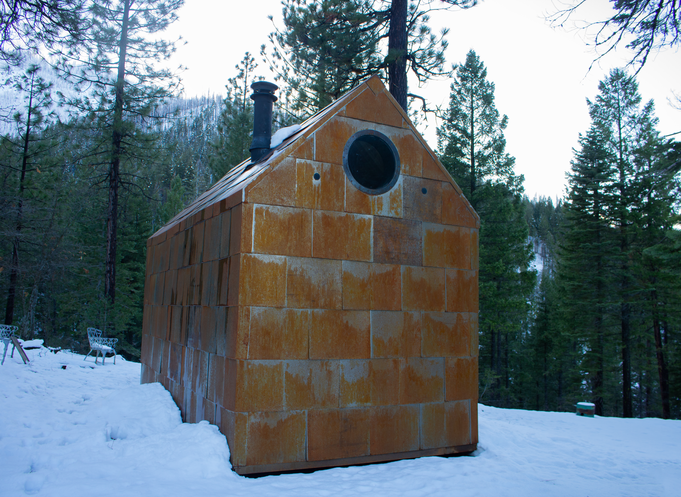 A small chapel made of orange tiles with a round window in a forested snowy landscape.
