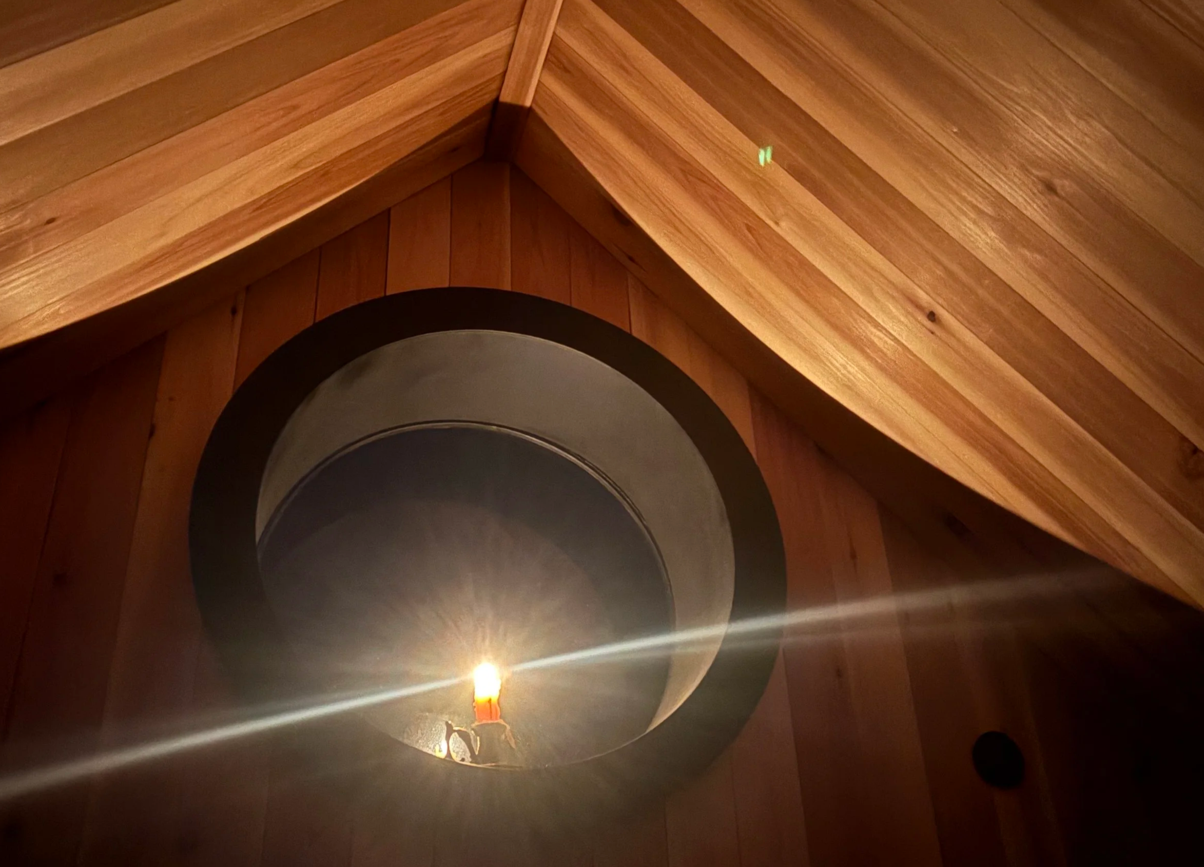 A candle burns in the interior of the Sauna, illuminating Western Red Cedar tongue and groove paneling and a pitch black porthole window.