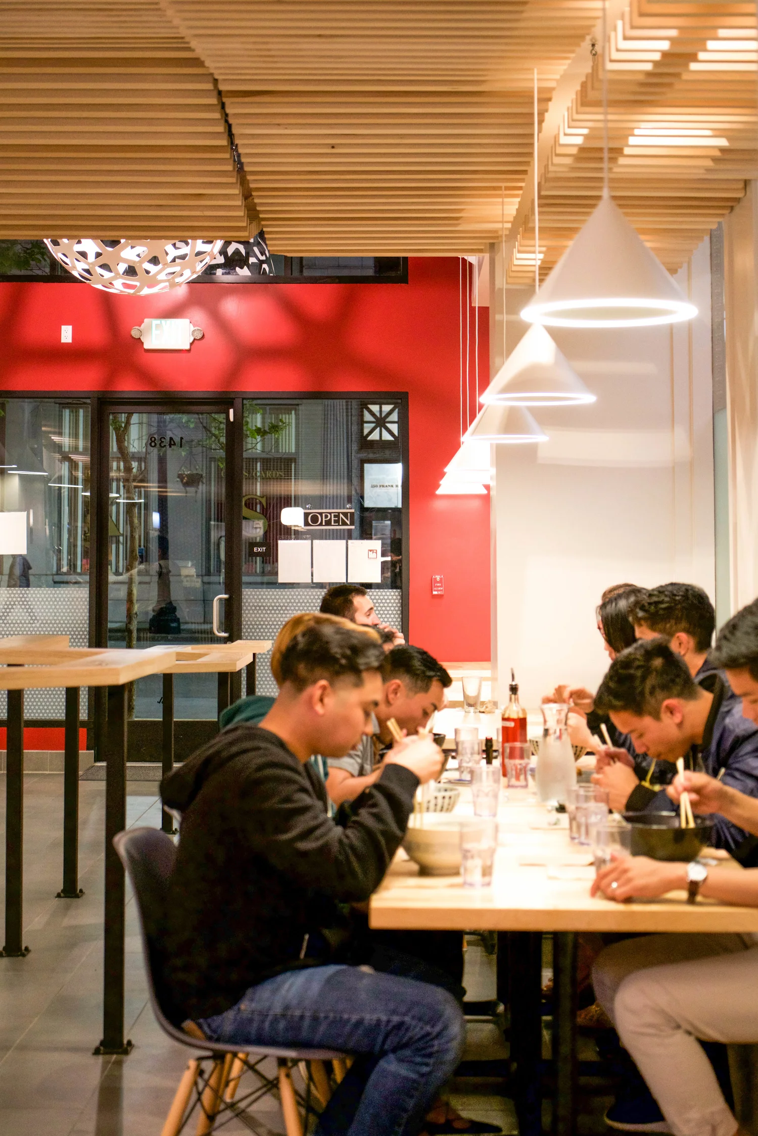 Patrons enjoy ramen at a long wooden table, surrounded by a well-lit restaurant, with wood and red accents.