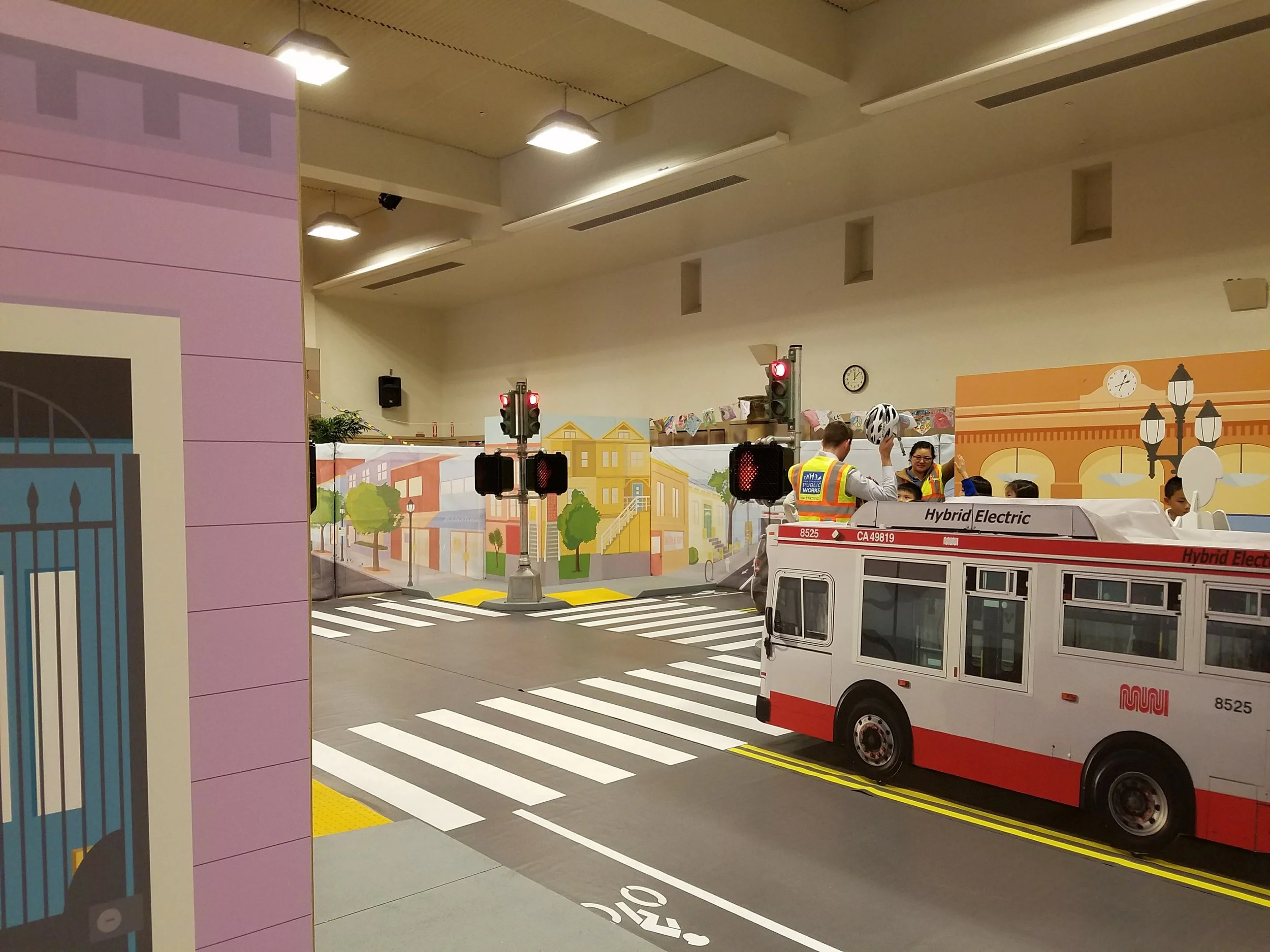 Indoor scene with a miniature city street display, including a toy bus labeled 'Hybrid Electric,' traffic lights, a crosswalk, and a background mural of buildings and trees. Two people are present; one is holding a bike helmet.