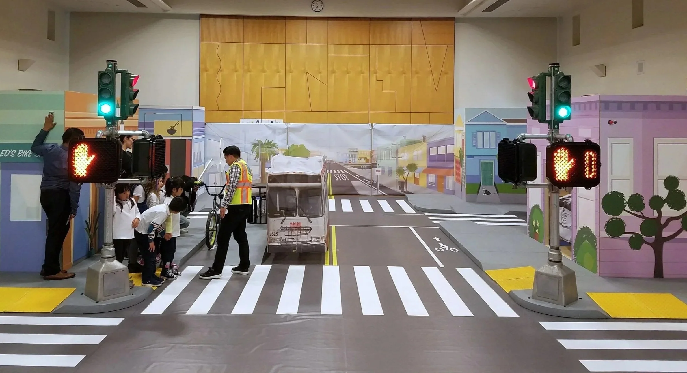 Indoor traffic safety exhibit with traffic lights, pedestrians, and a miniature city scene, including a cardboard bus, painted buildings, and crosswalks.