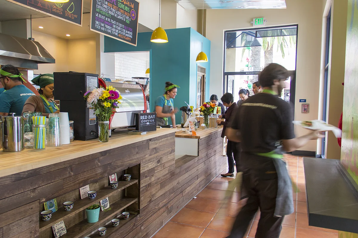 Inside a coffee shop with some employees and customers, a wooden counter with a flower vase, a coffee machine, and colorful cones on the counter. Customers are ordering, and staff are serving. Large windows with a view outside, showing palm trees and street reflections.