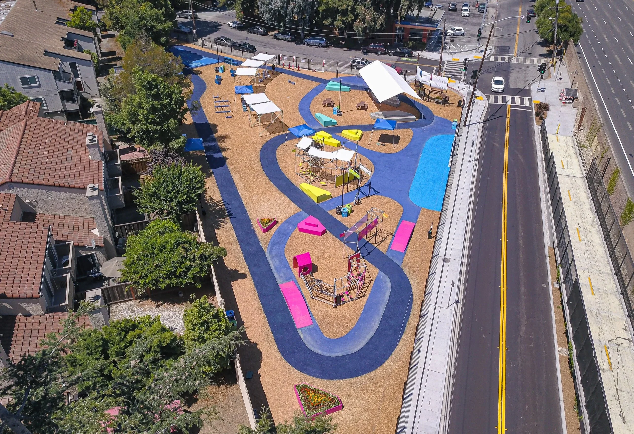 Aerial view of Boom Pop Park. The park has a series of winding pathways, seating areas, shade structures, and modular, brightly colored play structures. The park is nestled between a residential neighborhood and highway.