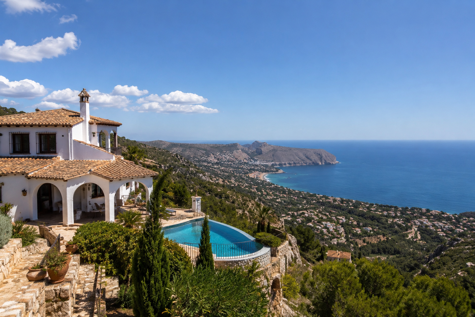 A white Mediterranean-style villa with stucco walls and tile roof on a hillside overlooking the ocean, with a curved swimming pool, lush green landscaping, and distant coast and hills under a blue sky with clouds.