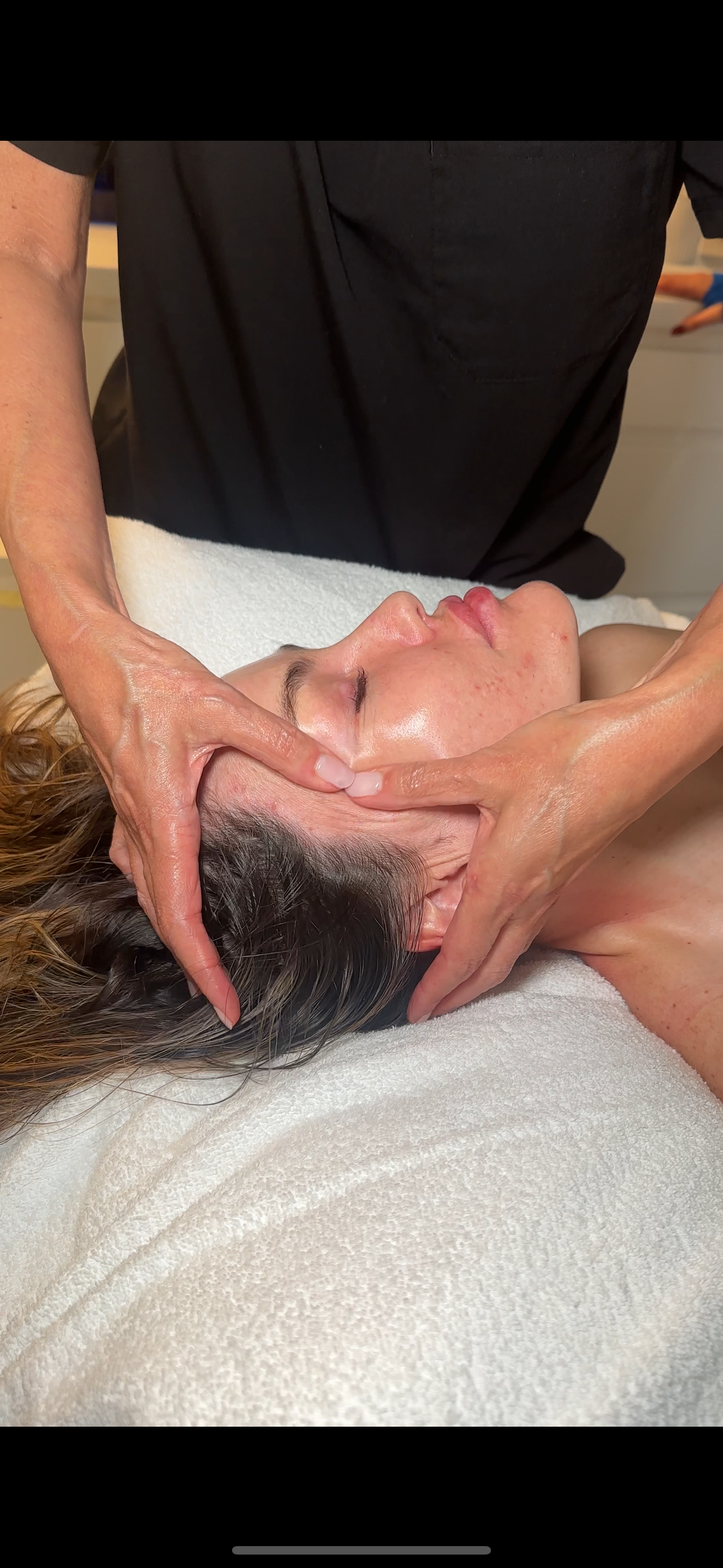 A person is receiving a facial massage while lying on a towel, with a massage therapist applying pressure to their face and head.