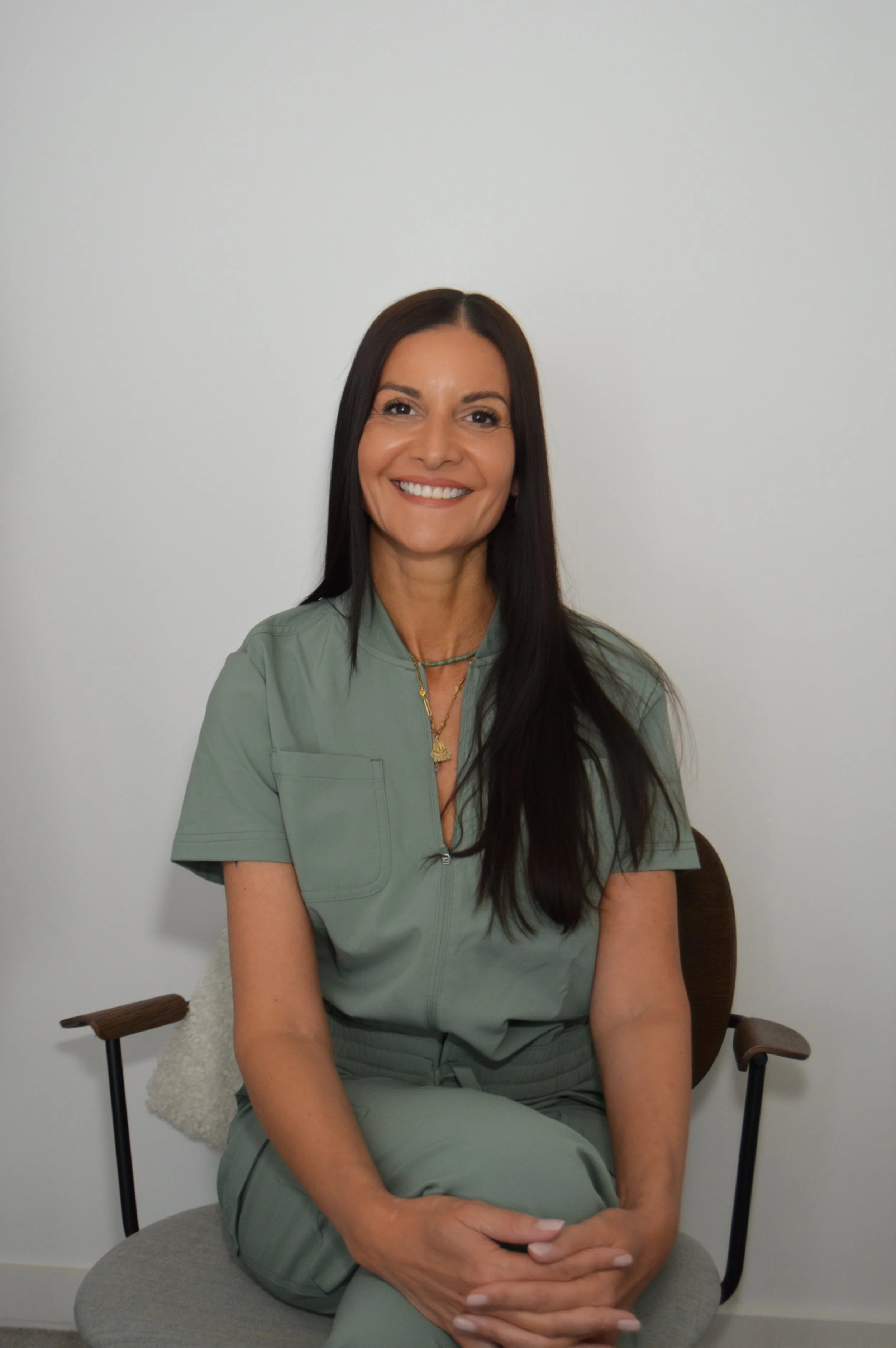 A woman with long dark hair smiling, sitting on a gray cushioned stool against a plain white wall. She is wearing green scrubs and sitting with her hands resting on her lap.