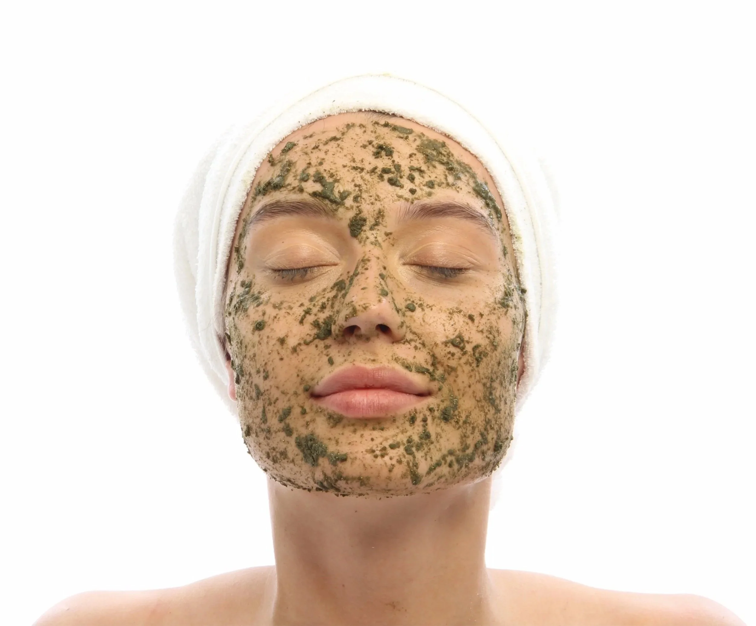 Person with closed eyes and a white towel wrapped around the head, applying a green facial mask with visible patches over the face and forehead.