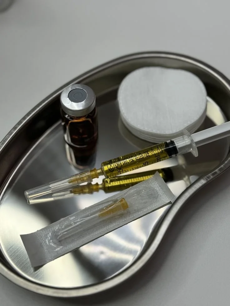 A metal tray containing a small brown medicine bottle, a stack of white cotton pads, a syringe with yellow liquid, a sealed sterile needle, and a small vial.