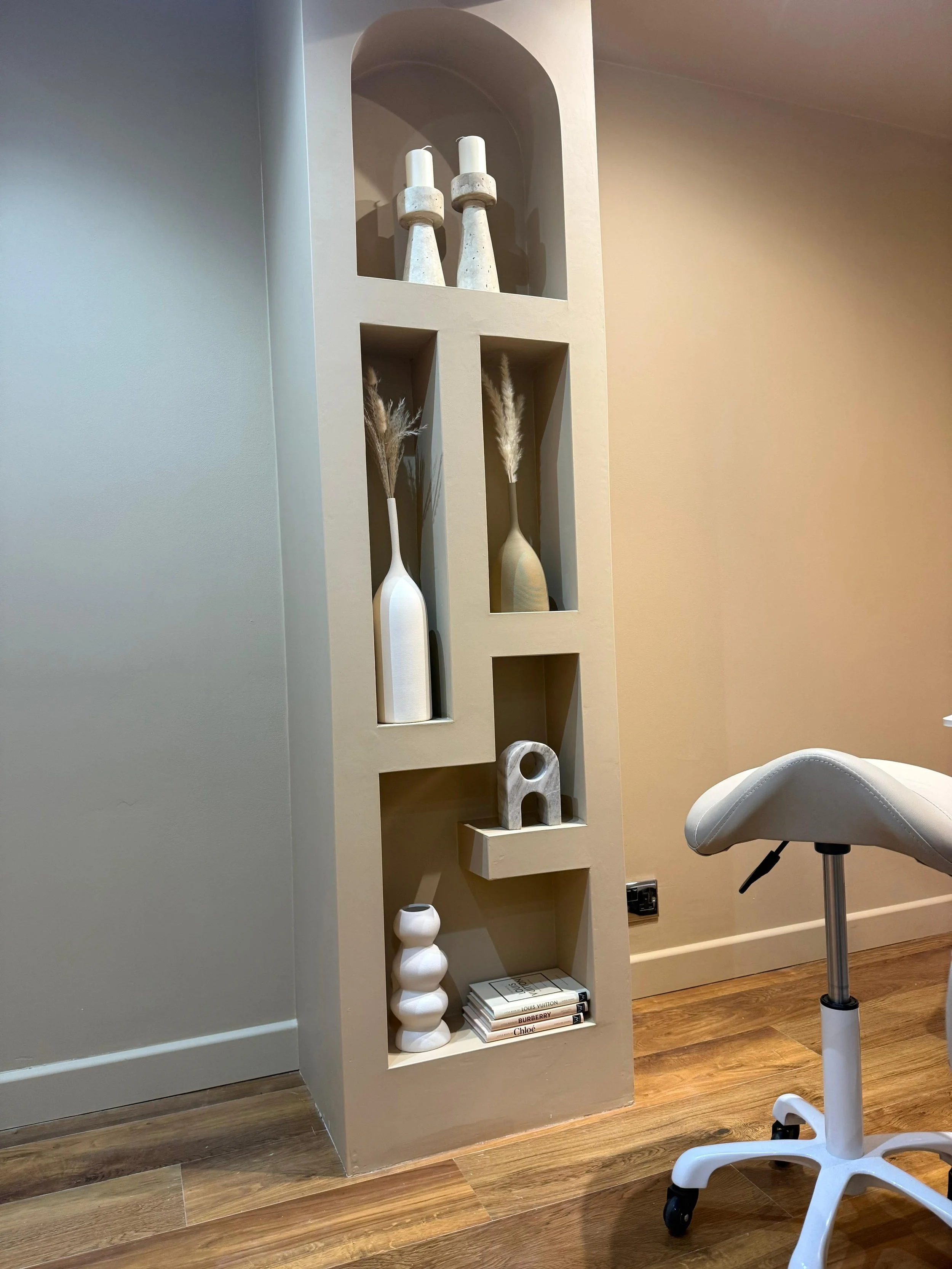 A beige wall with built-in wall niches containing decorative vases and candles, next to a white desk chair on a wooden floor.