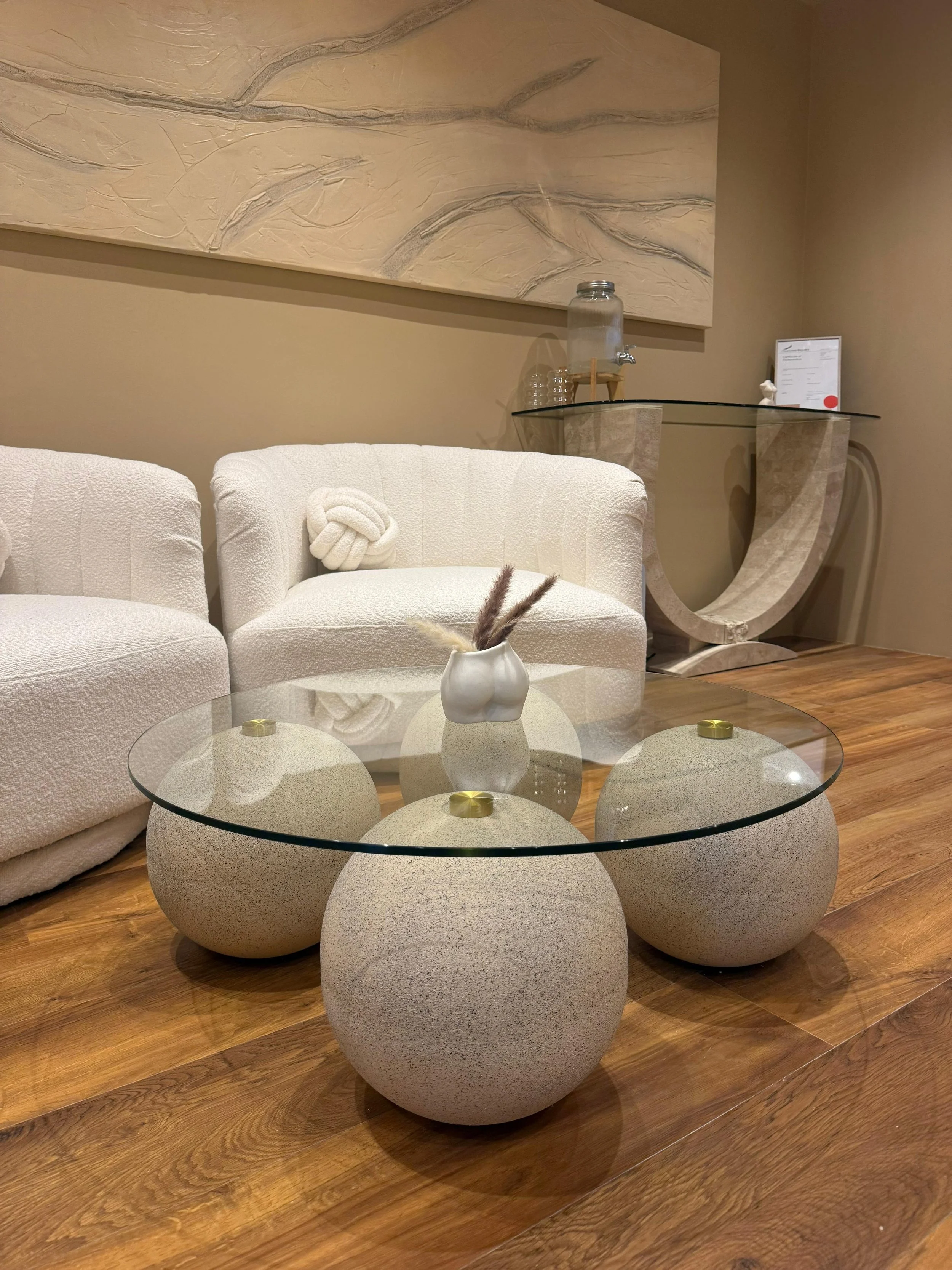 Contemporary living room with white textured sofas, a glass-topped coffee table with textured stone bases, a wall-mounted abstract textured artwork, a curved stone console table with a water dispenser, glass cups, and a framed certificate.