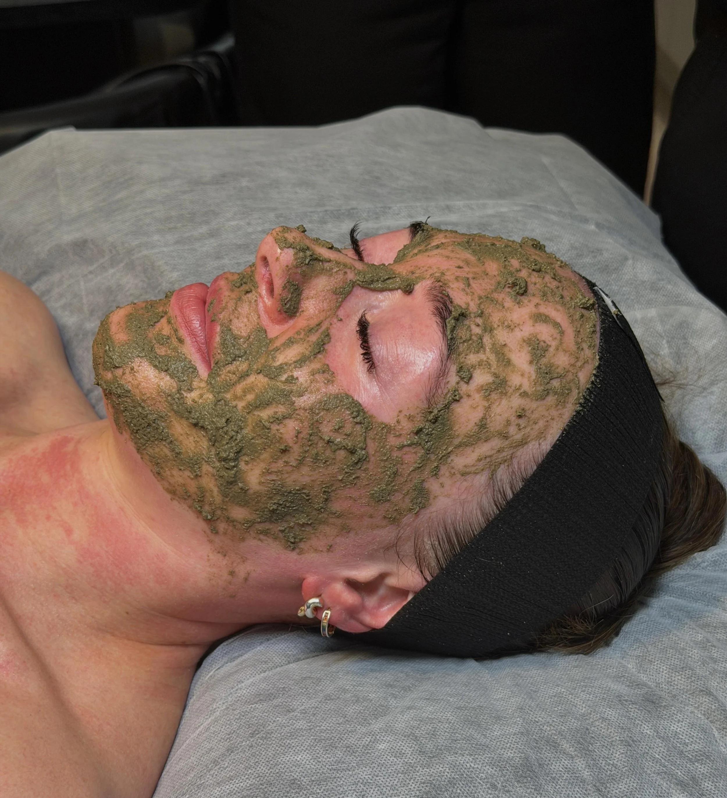 Close-up of a person's face with green facial mask applied, lying in a spa or skincare treatment setting.