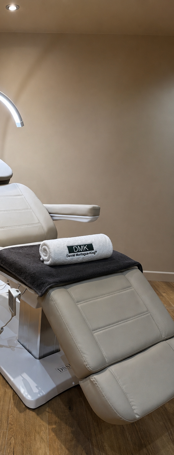 Medical examination chair in a clinic or spa, with a rolled towel marked 'DMK Danné Montague-King' placed on a blanket.
