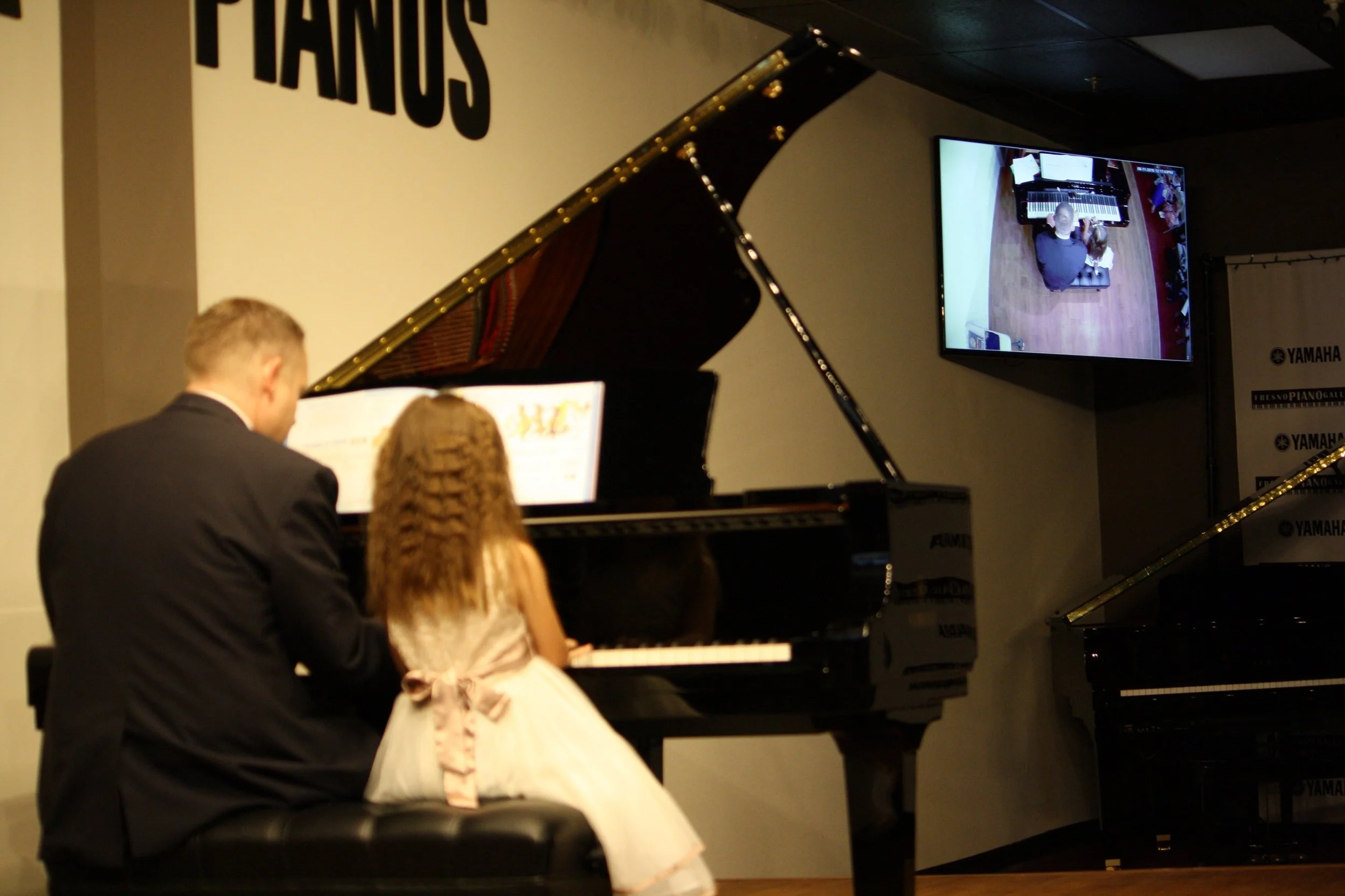 A piano recital featuring a grand piano is being played by a person dressed in a black suit, with a young girl in a white dress holding sheet music nearby.