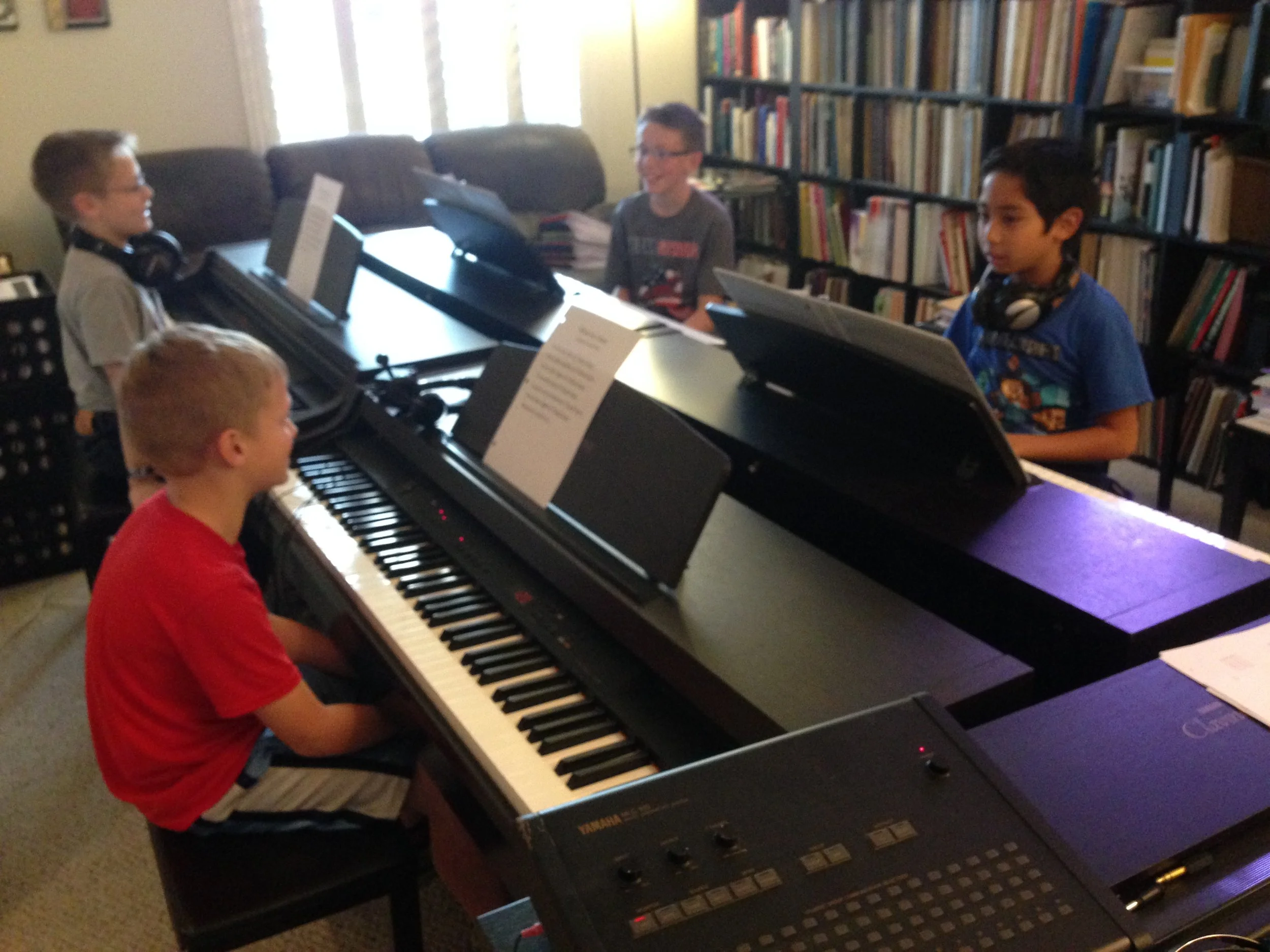 Four children are in a piano lab, with four boys sitting at electric pianos. They are in front of a large bookshelf filled with books.
