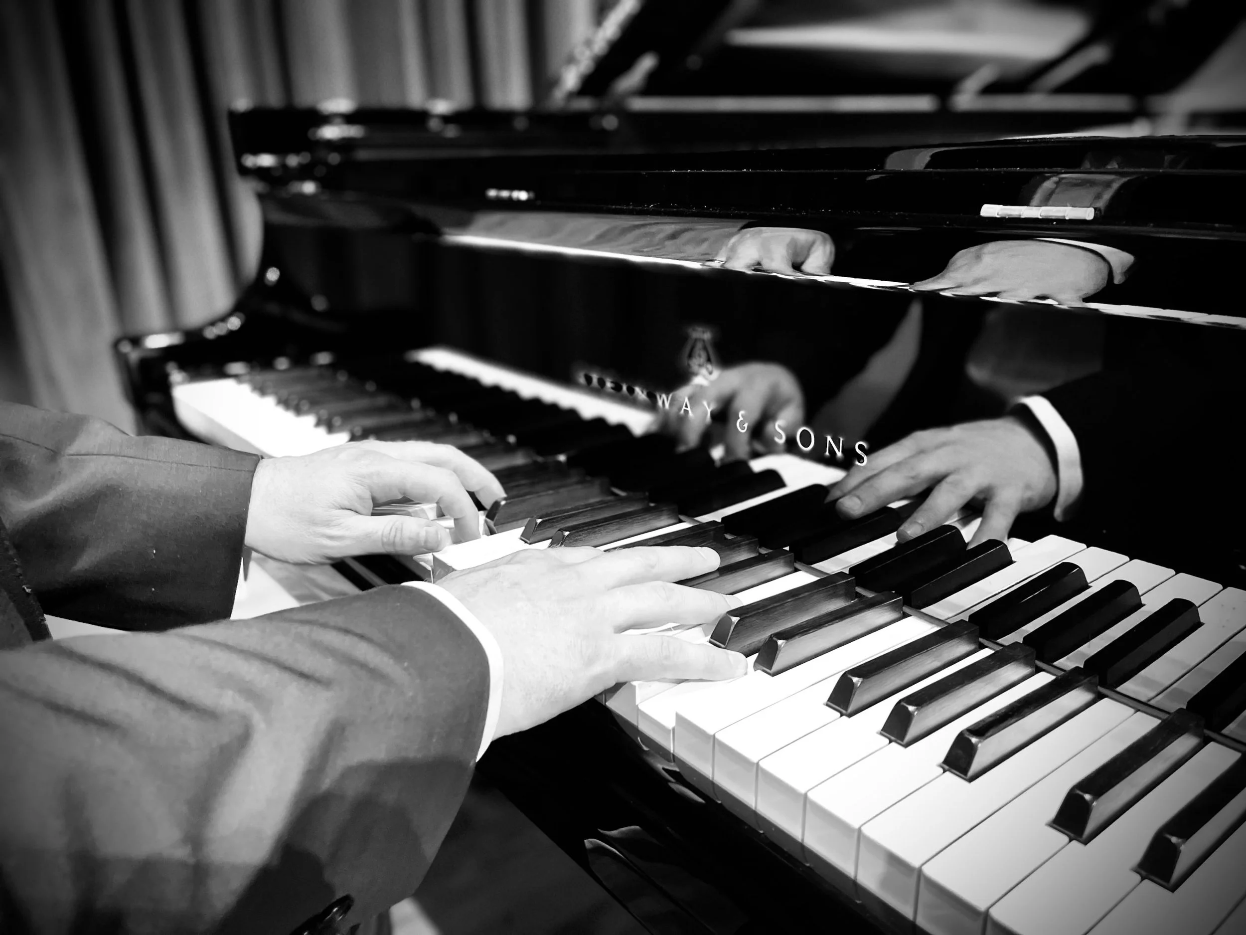 Black and white photo of someone playing the piano with the reflection of their hands showing in the fall board. 