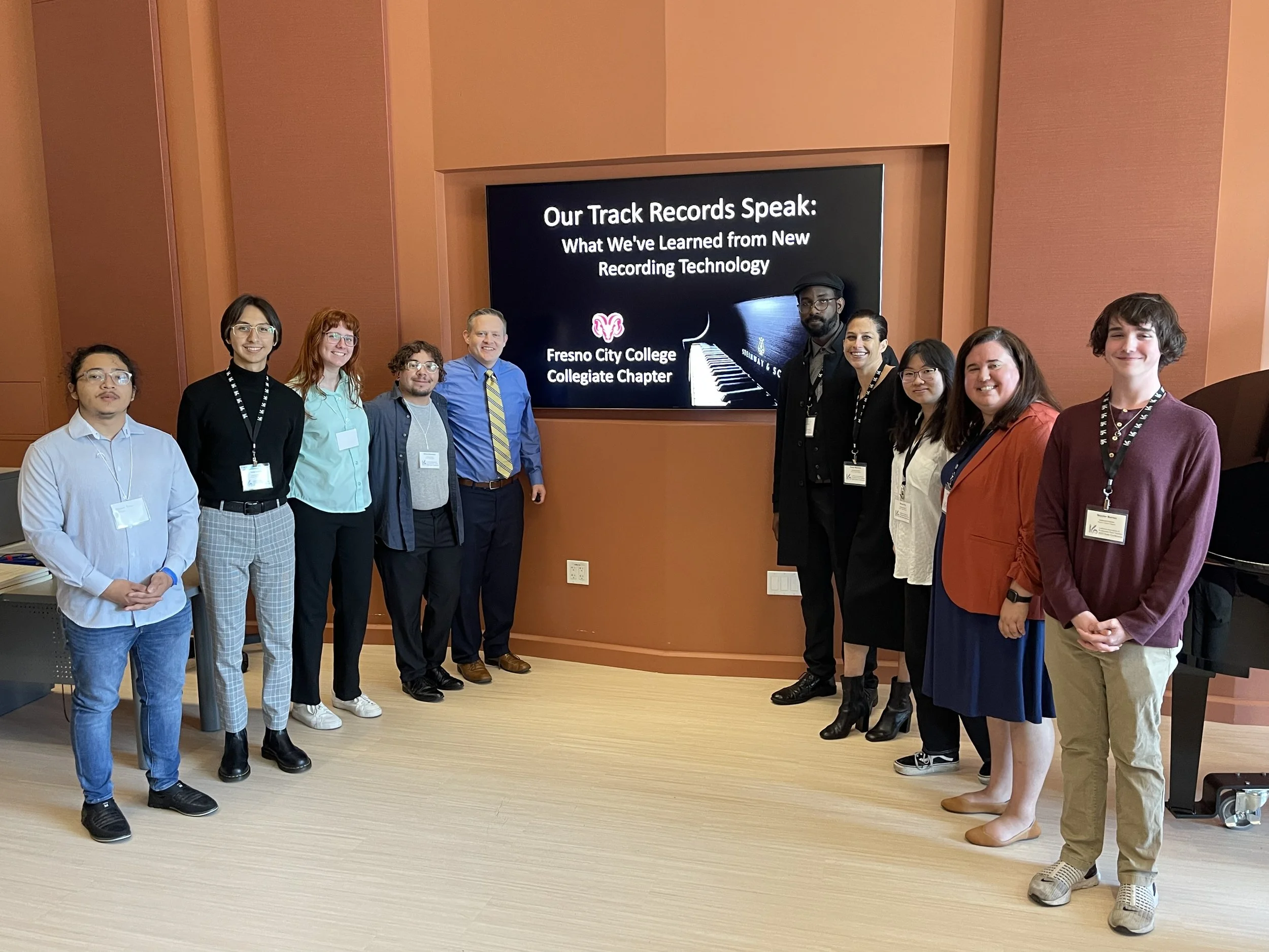 Group of ten people standing in front of a large TV screen that displays 'Our Track Records Speak: What We've Learned from New Recording Technology' and 'Fresno City College Collegiate Chapter.' The group is diverse, dressed in casual to business casual attire, smiling for the photo inside a room with reddish-brown walls and a light wood floor.