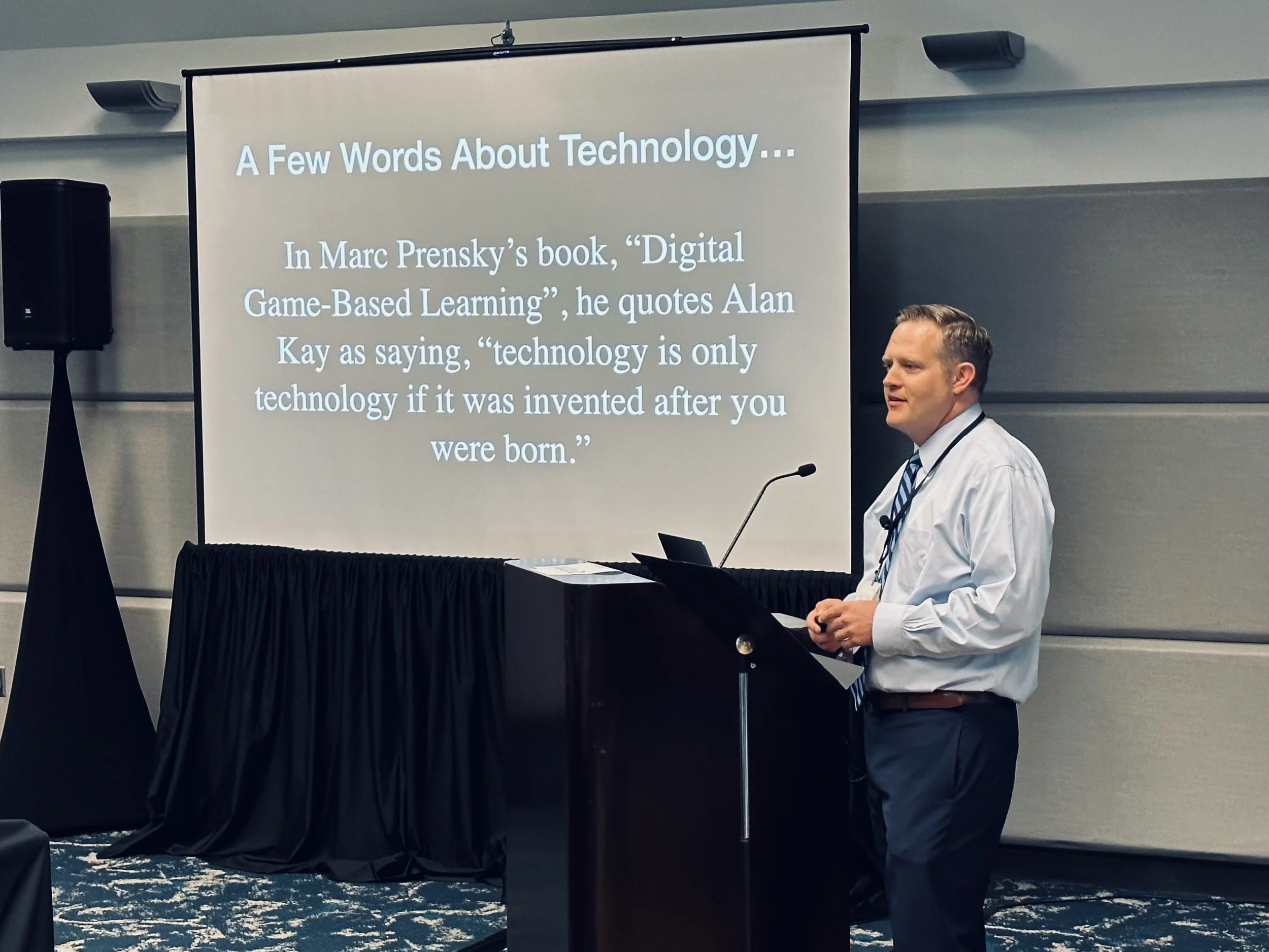A man wearing a white shirt and striped tie standing next to a podium during a presentation. A large screen displays a slide titled 'A Few Words About Technology...' with text quoting Marco Prensky's book about digital game-based learning.