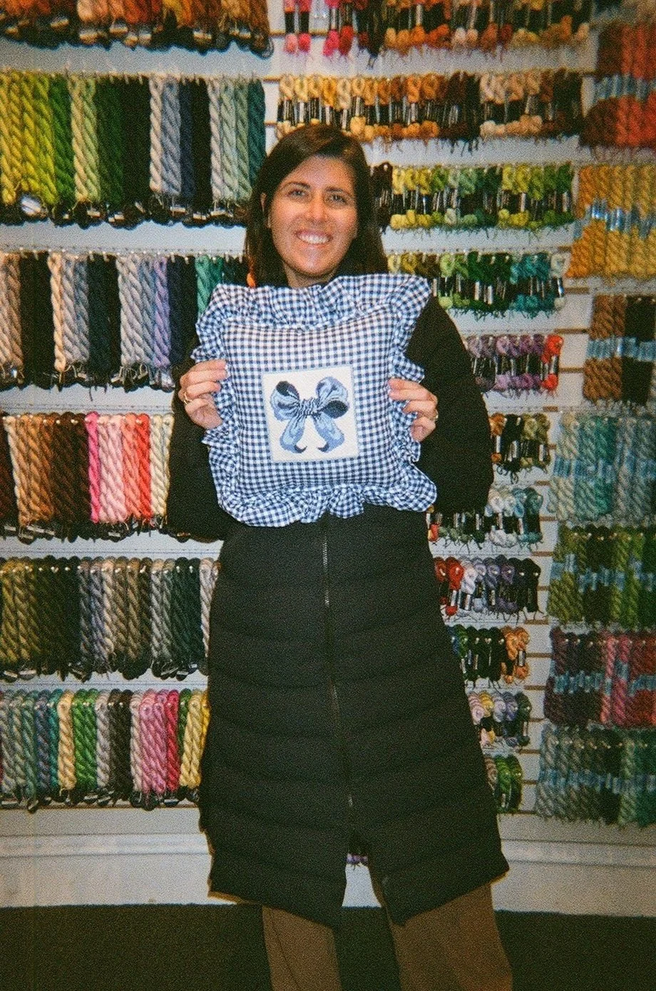 A woman smiling and holding a checkered pillow with a bow design in front of a wall full of colorful yarns.