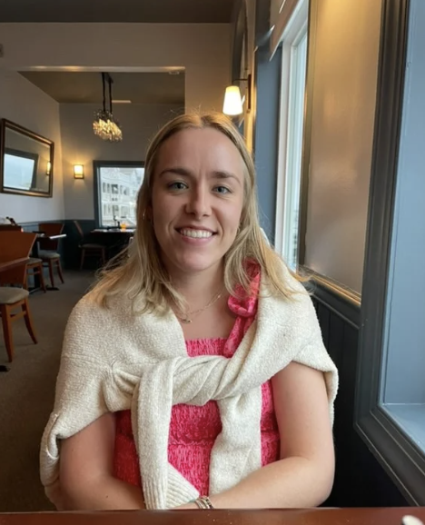 A young woman with blonde hair smiling in a cafe, wearing a pink top with a cream sweater draped over her shoulders, sitting near a window.