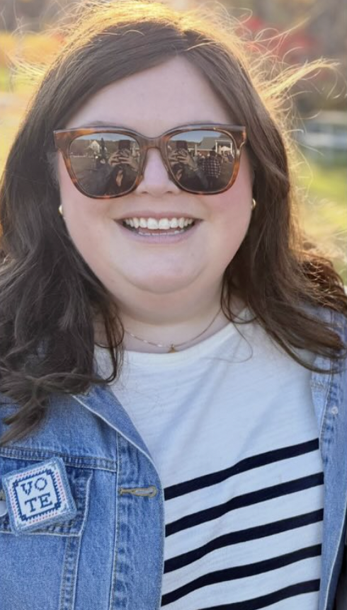 A woman smiling outdoors, wearing large sunglasses, a striped shirt, and a denim jacket with a "VOTE" patch, during sunset.