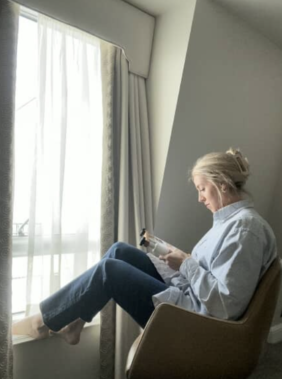 A woman with blonde hair in a bun sitting in a chair near a large window with sheer curtains, reading a magazine and listening to earphones.