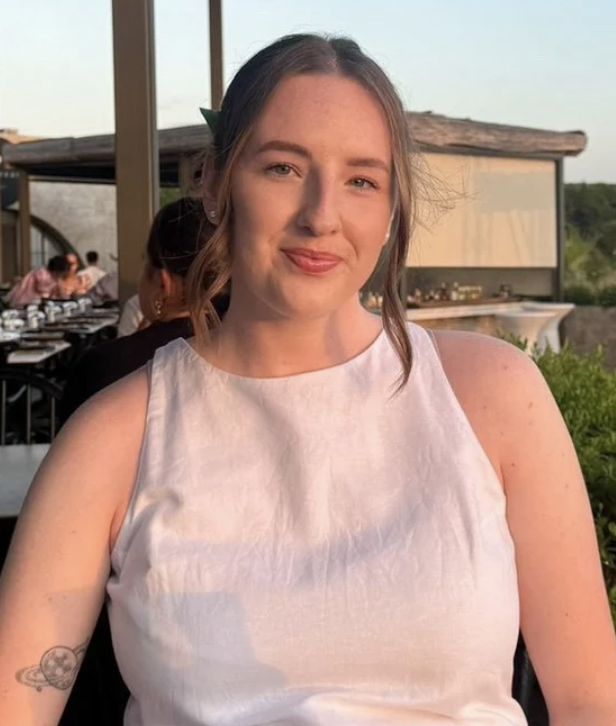 A young woman with light brown hair and a slight smile, wearing a sleeveless white top, seated outdoors during the daytime with a blurred background of other people and greenery.