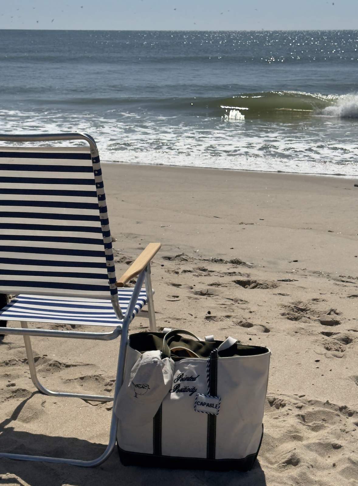 Empty beach chair next to a white tote bag with a sea turtle logo, on sandy beach near ocean waves.