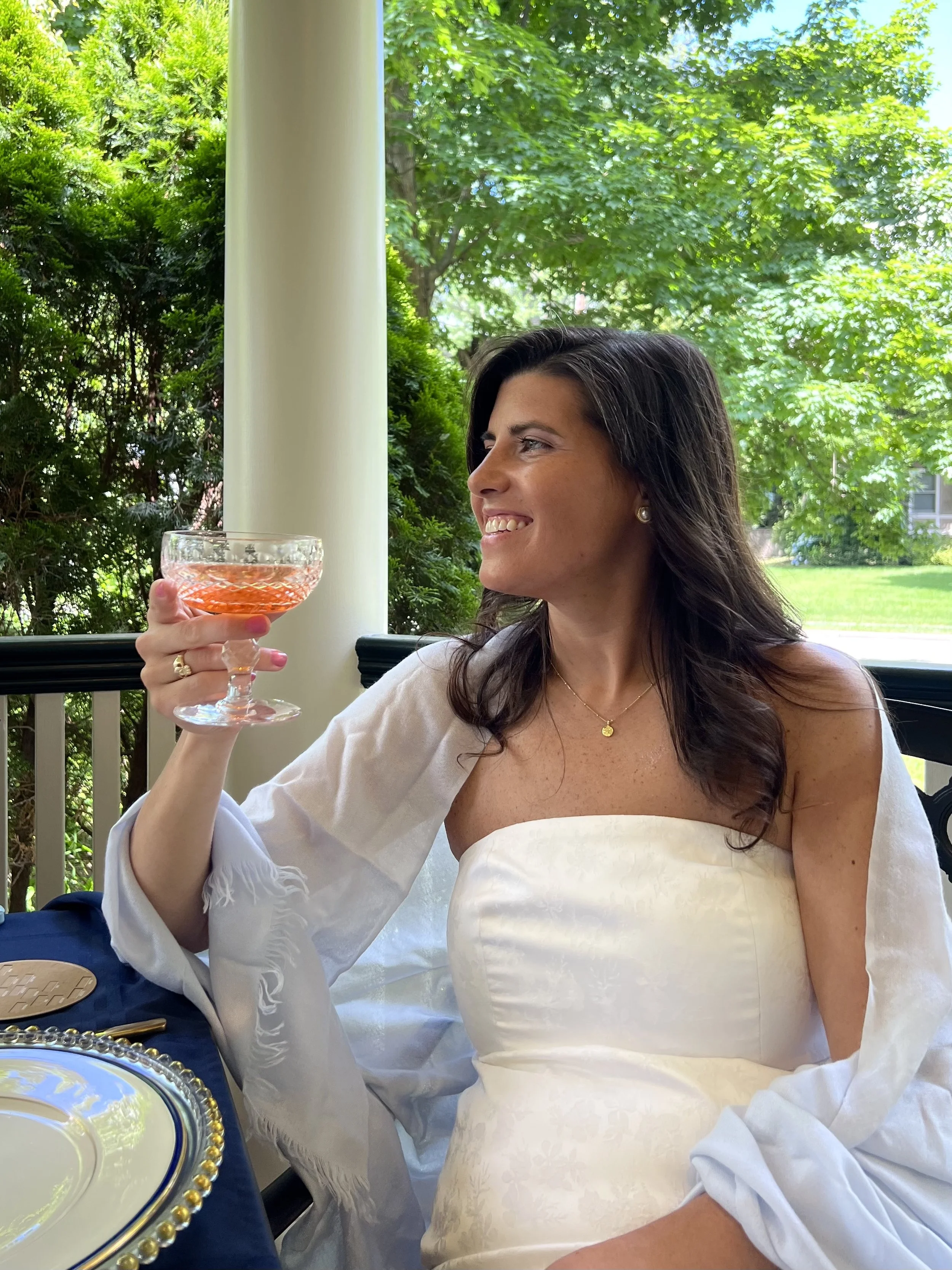 A woman with dark hair and light skin, wearing a white strapless dress and a white shawl, is sitting at a table outside on a porch, holding a glass of pink beverage with a smile, with green trees and a grassy yard in the background.