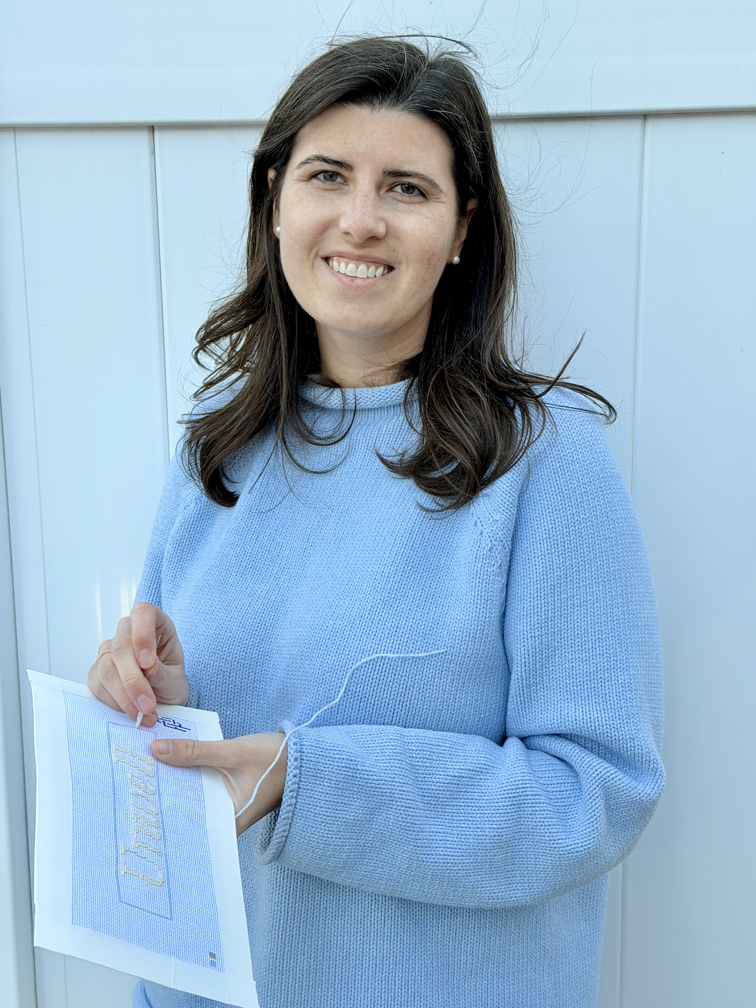 A woman with dark brown hair, wearing a light blue sweater, is holding an embroidery pattern while standing against a light blue wall.