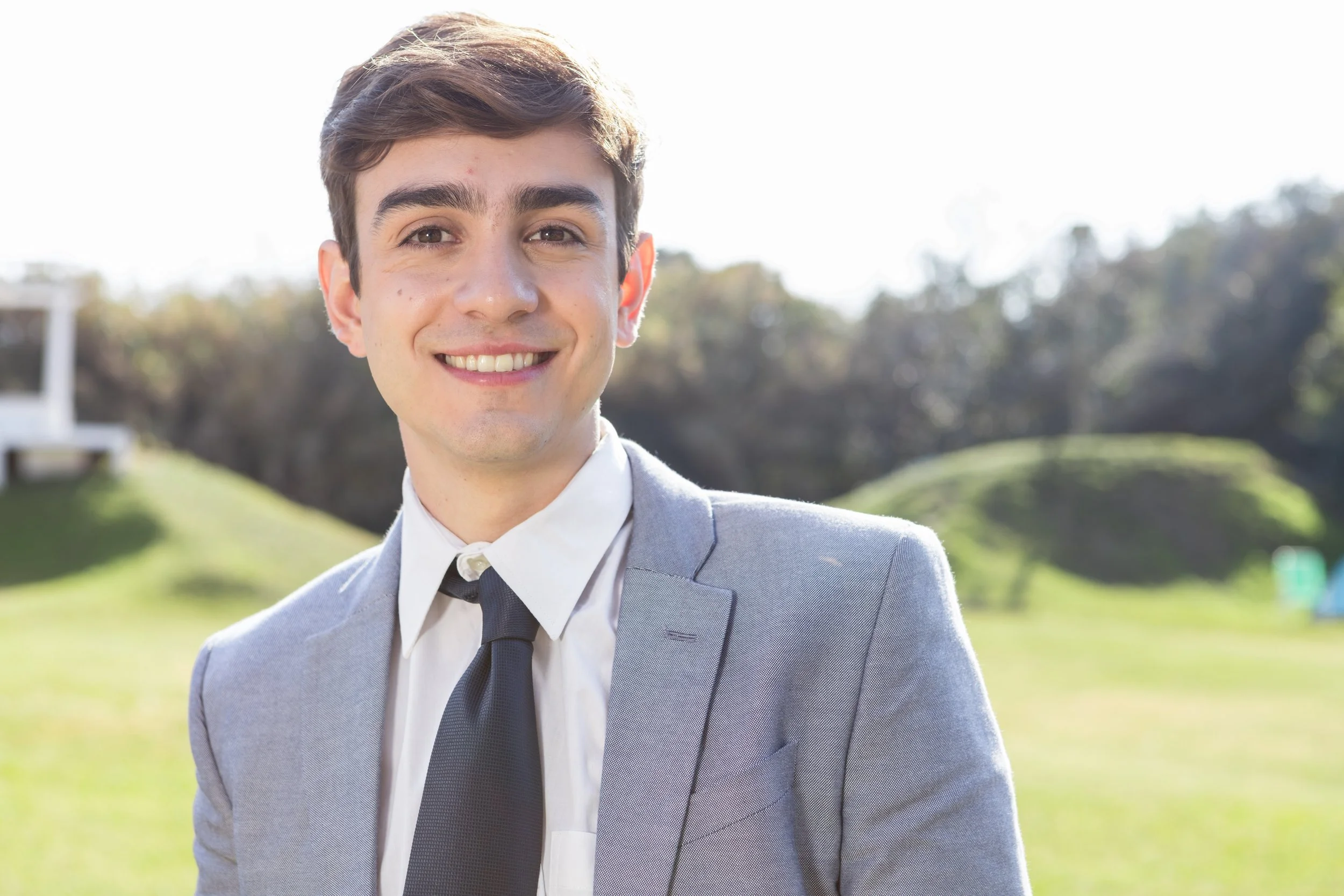 Young man in a gray suit and black tie smiling outdoors on a sunny day.