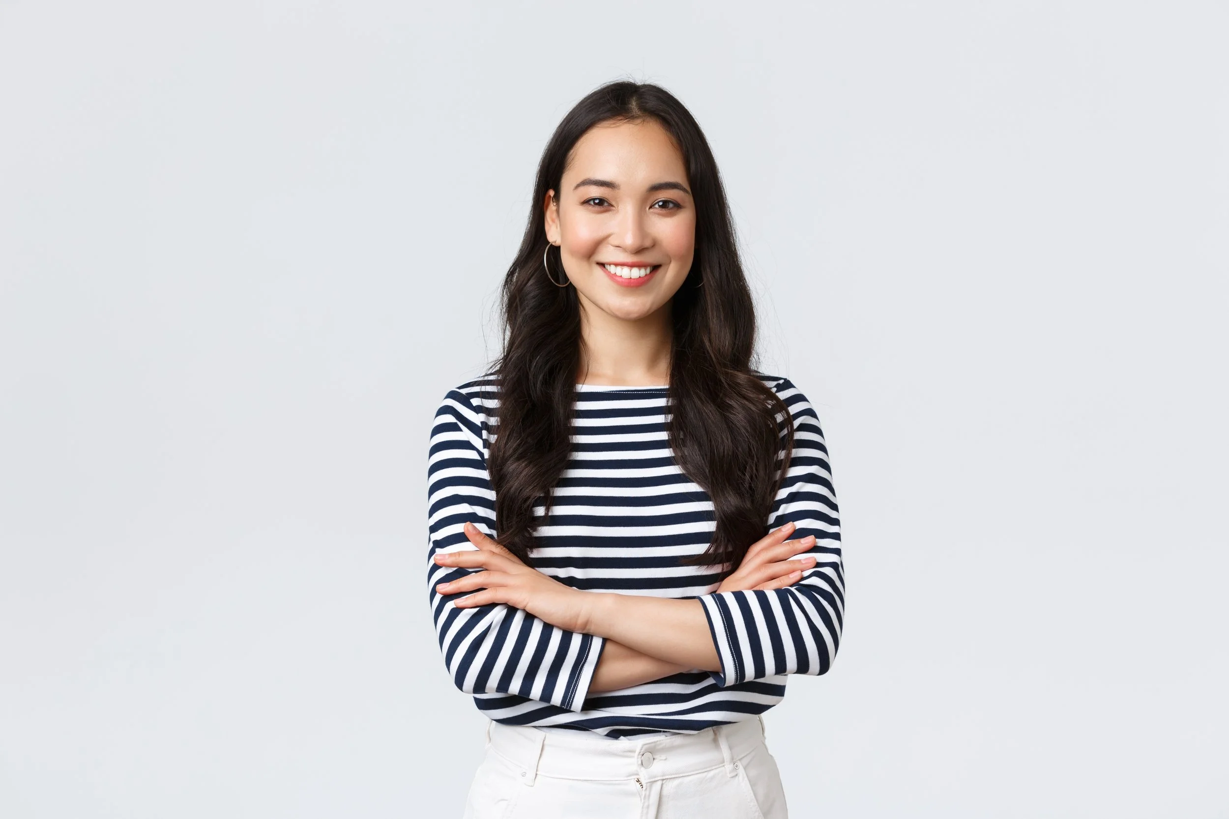 A young woman with long dark hair, smiling, wearing a navy and white striped shirt and white pants, standing with crossed arms against a plain light background.
