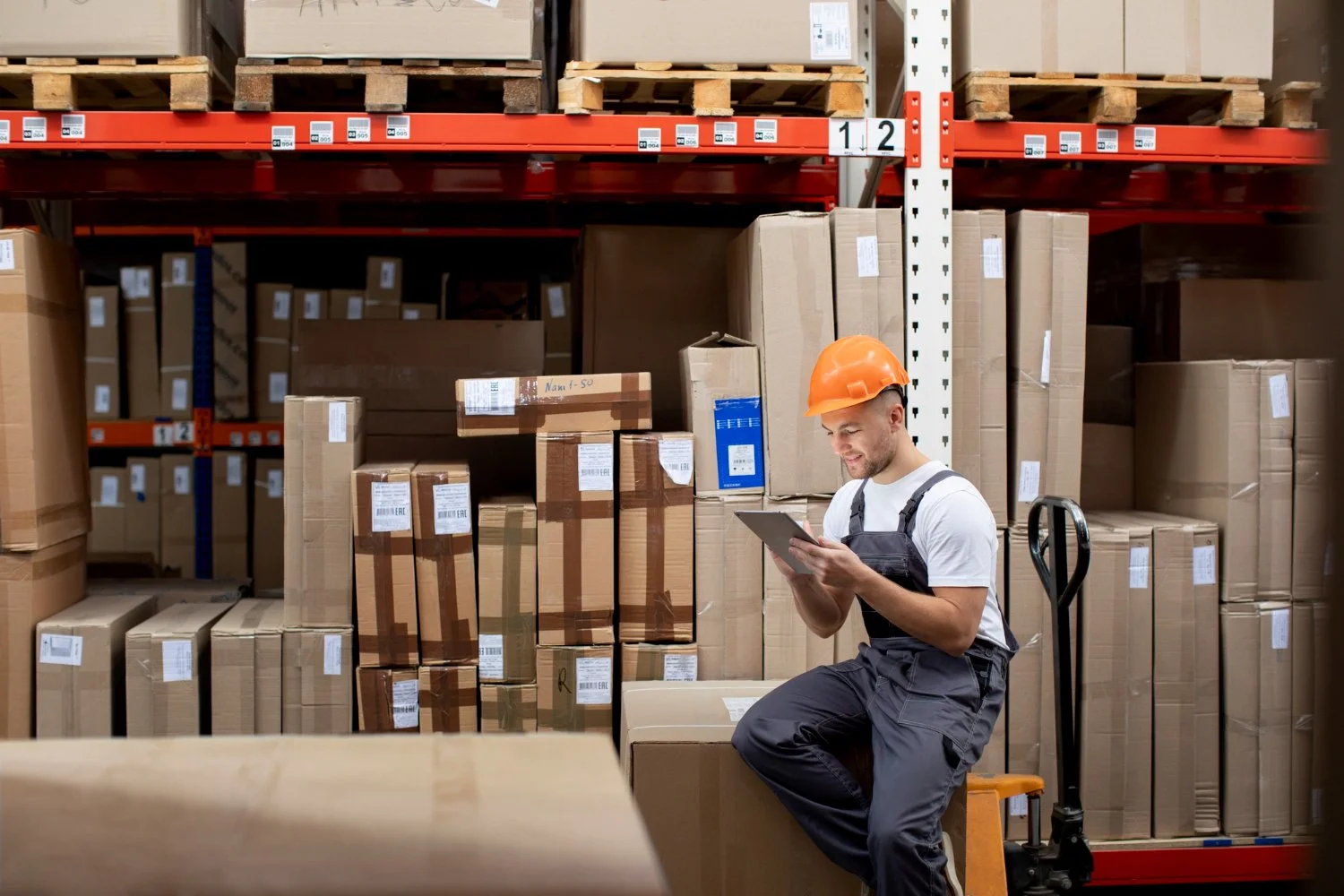 A worker with an orange safety helmet sitting on a stool in a warehouse, looking at a tablet. The warehouse is filled with shelves of cardboard boxes and pallets.