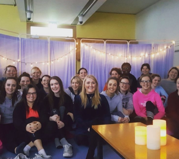 Group of young women sitting together indoors, smiling for a photo, with purple curtains and string lights in the background, and candles on a table in the foreground.