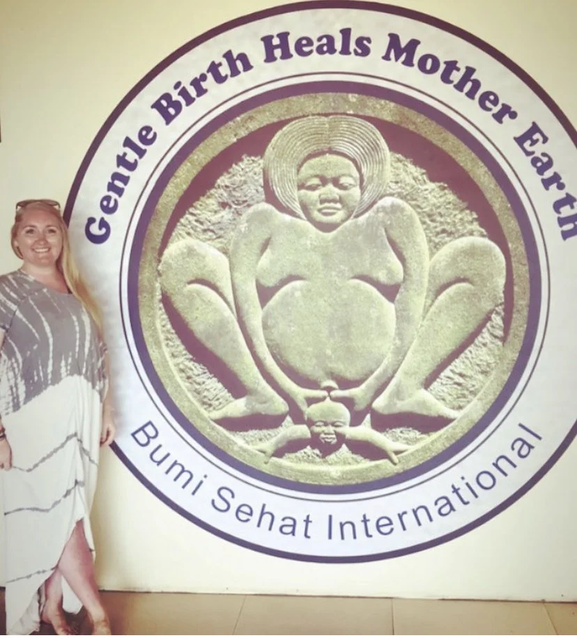 A woman standing next to a large circular logo with a sandy sculpture of a mother and child at its center. The text around the logo reads 'Gentle Birth Heals Mother Earth' and 'Bumi Sehat International'.