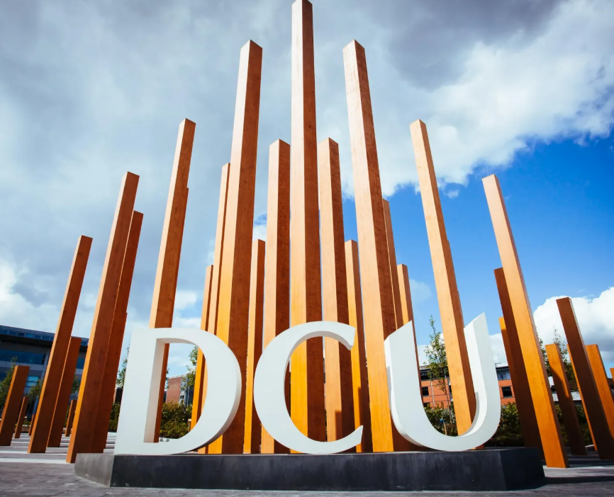 Large white letters spelling 'DCU' in front of tall wooden columns outdoors under a partly cloudy sky.