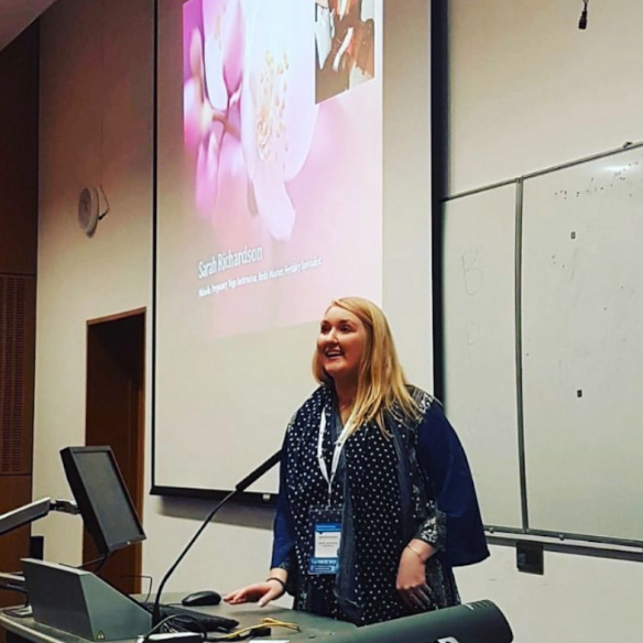 A woman with blonde hair standing at a podium in a classroom or conference room, smiling. Behind her is a large projection screen displaying a presentation slide and a whiteboard.