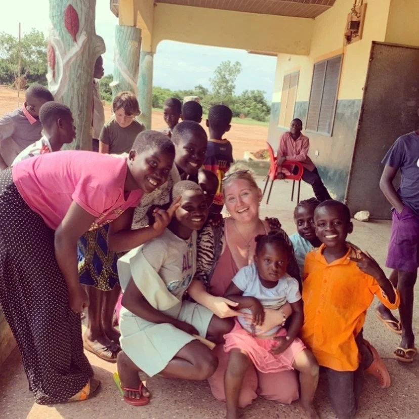 A group of children and a young woman pose smiling outdoors on a porch, with some children sitting and others standing around her.